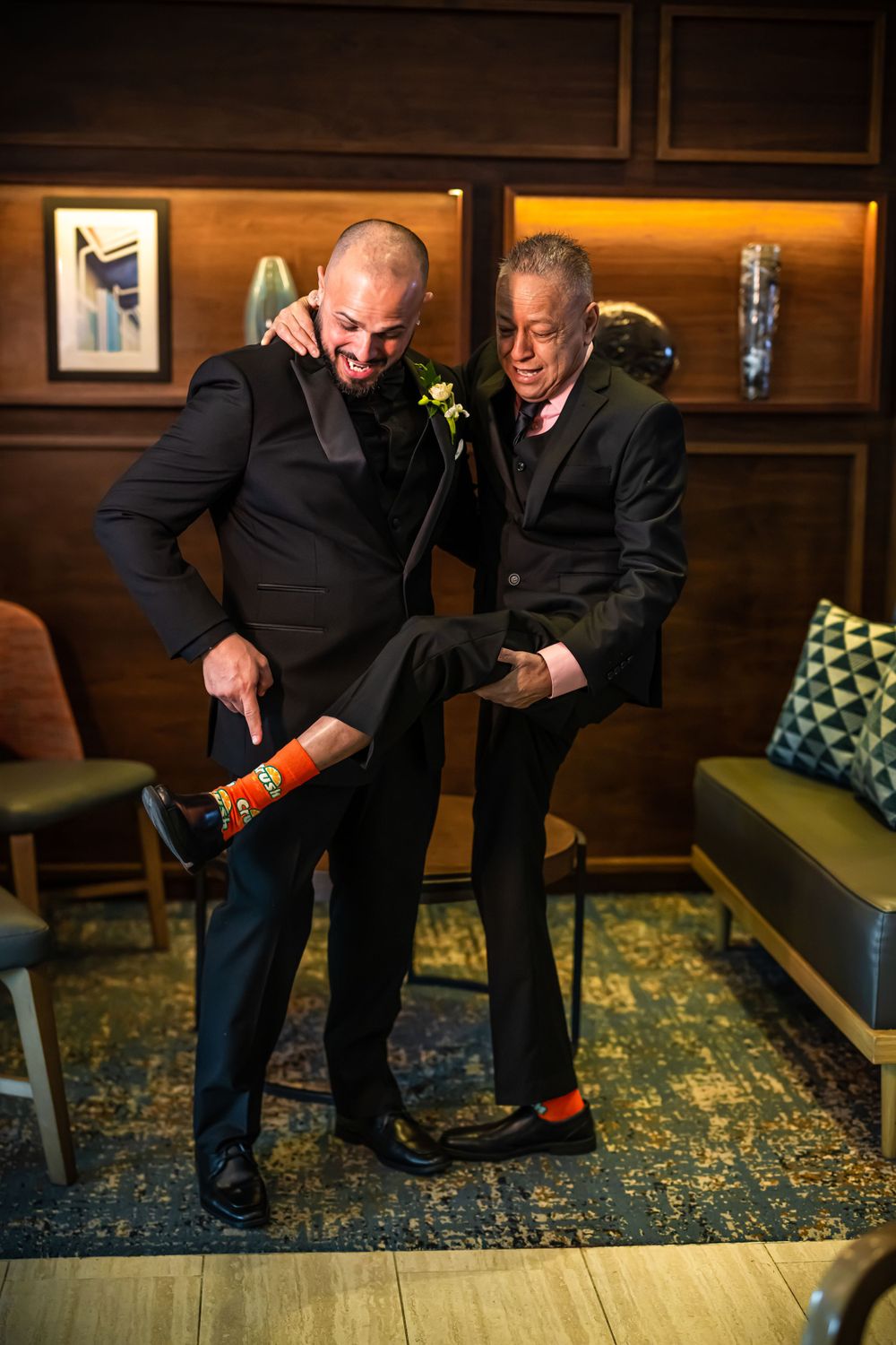 Two people in suits show off colorful socks while posing playfully on a patterned rug.
