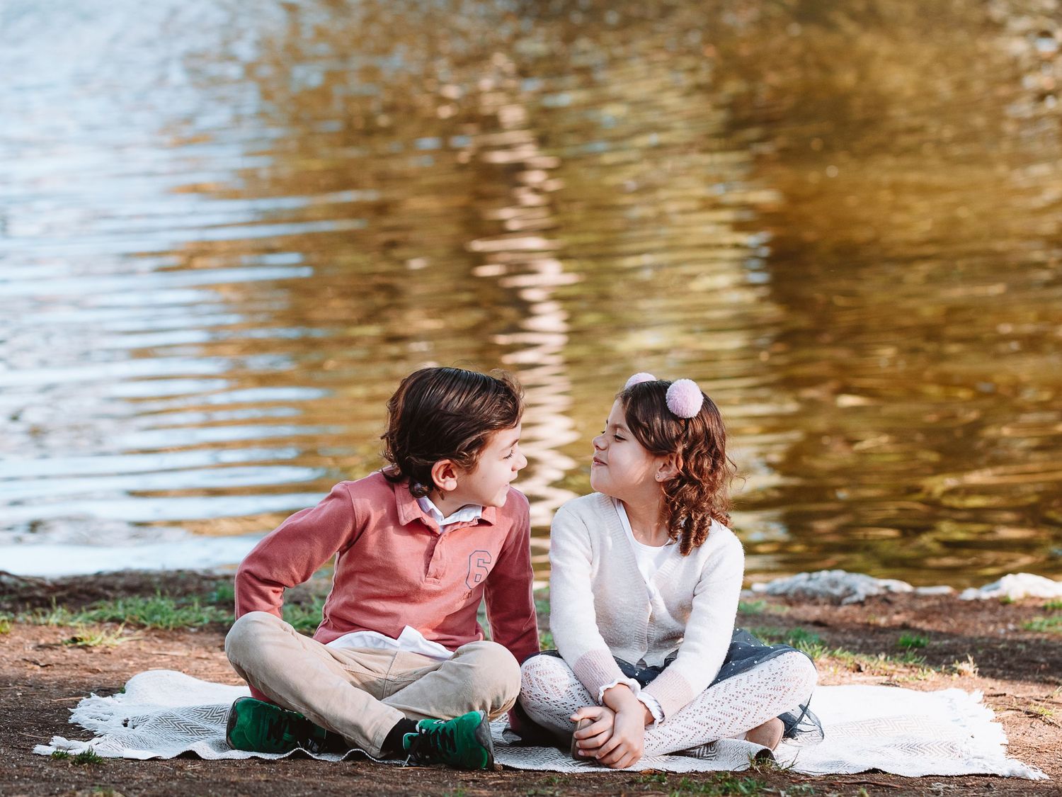 A couple sits together on a blanket by a lake with golden reflections in the water.