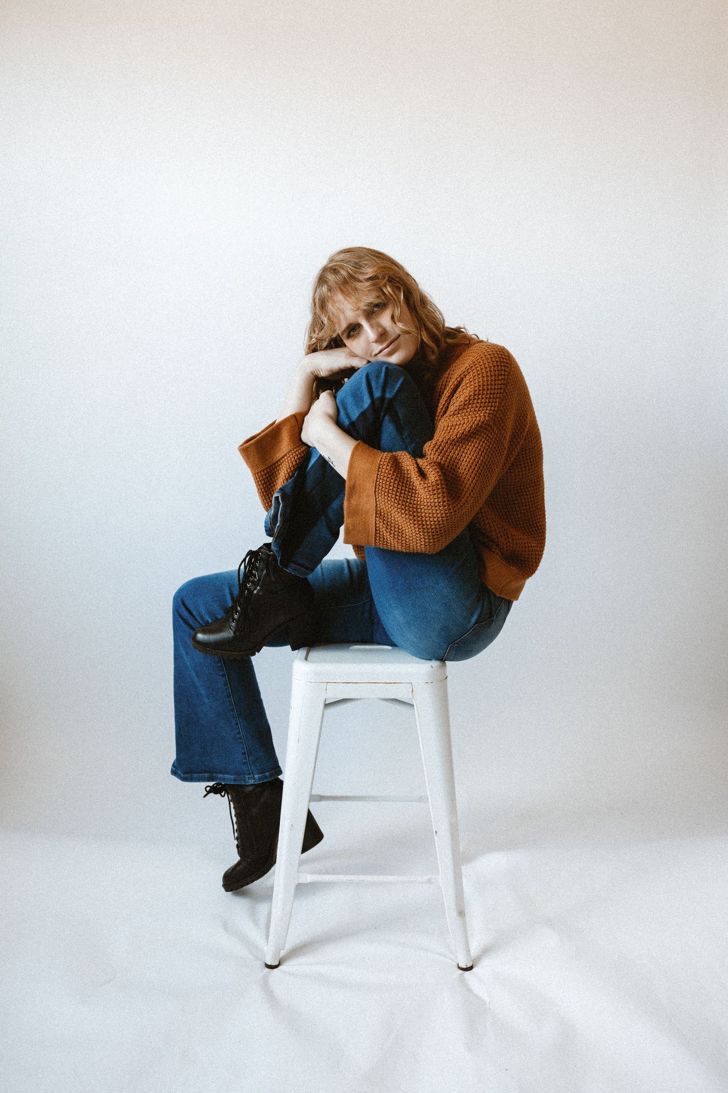 A person in a brown chunky sweater and blue jeans sits curled up on a white stool against a neutral background.