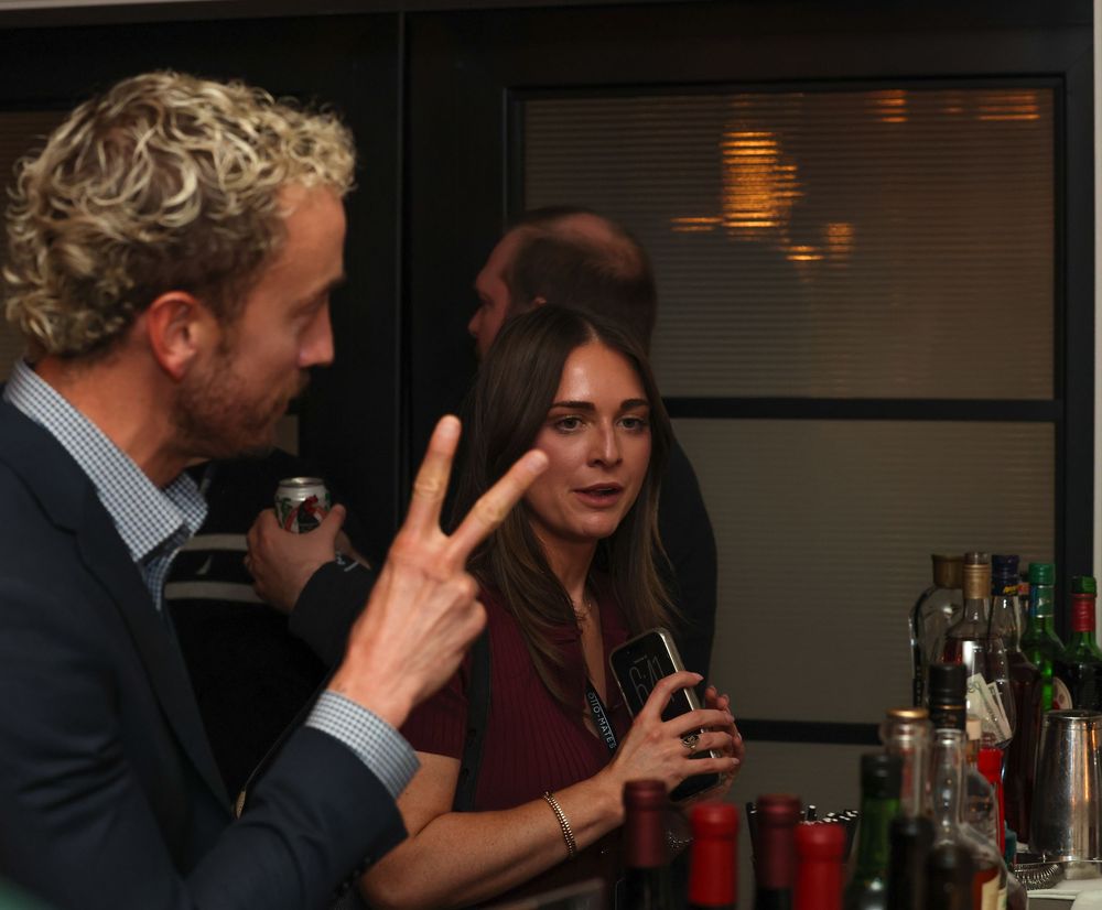 Two people having a conversation at a bar with drinks and bottles visible in the background.