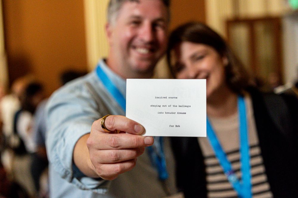 Two people wearing blue lanyards hold up a small card at a professional event.