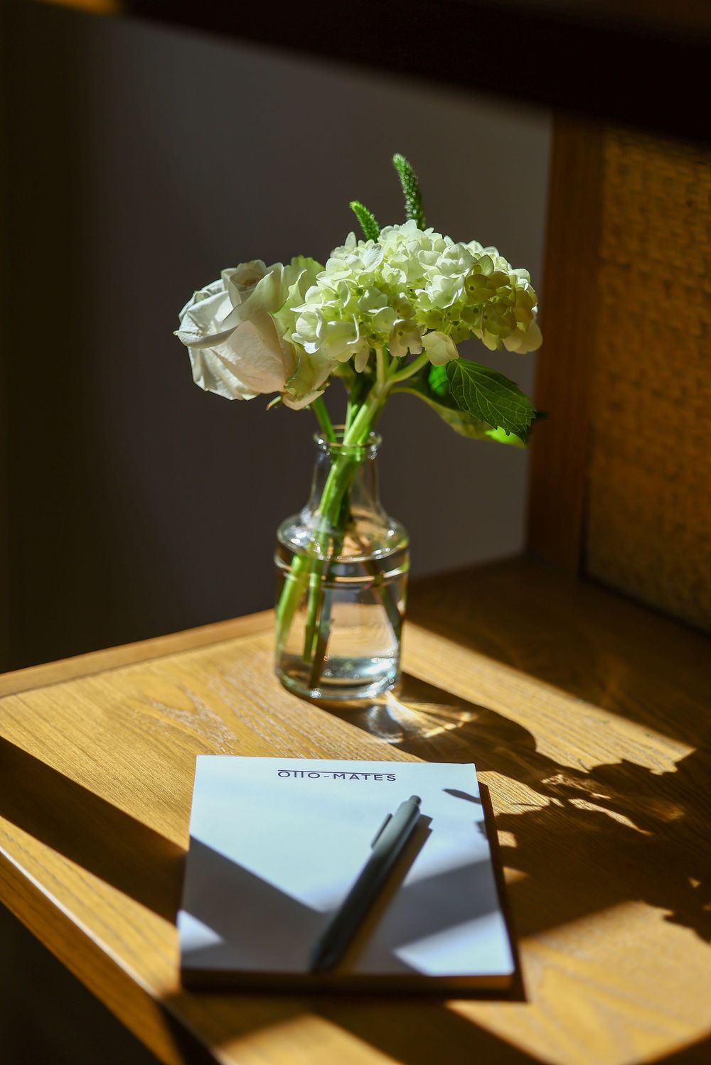 White flowers in a glass vase cast dramatic shadows on a wooden table with notebook and pen in natural sunlight.