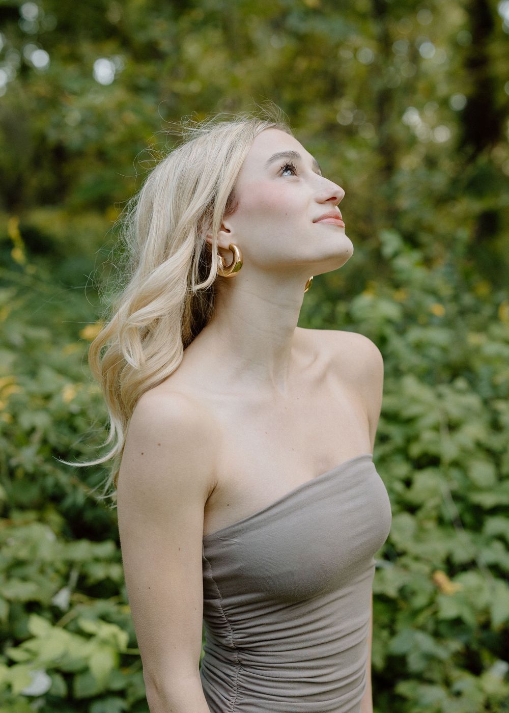 Blonde woman in strapless gray dress with gold hoop earrings gazing upward, surrounded by lush green foliage in Sacramento, California.