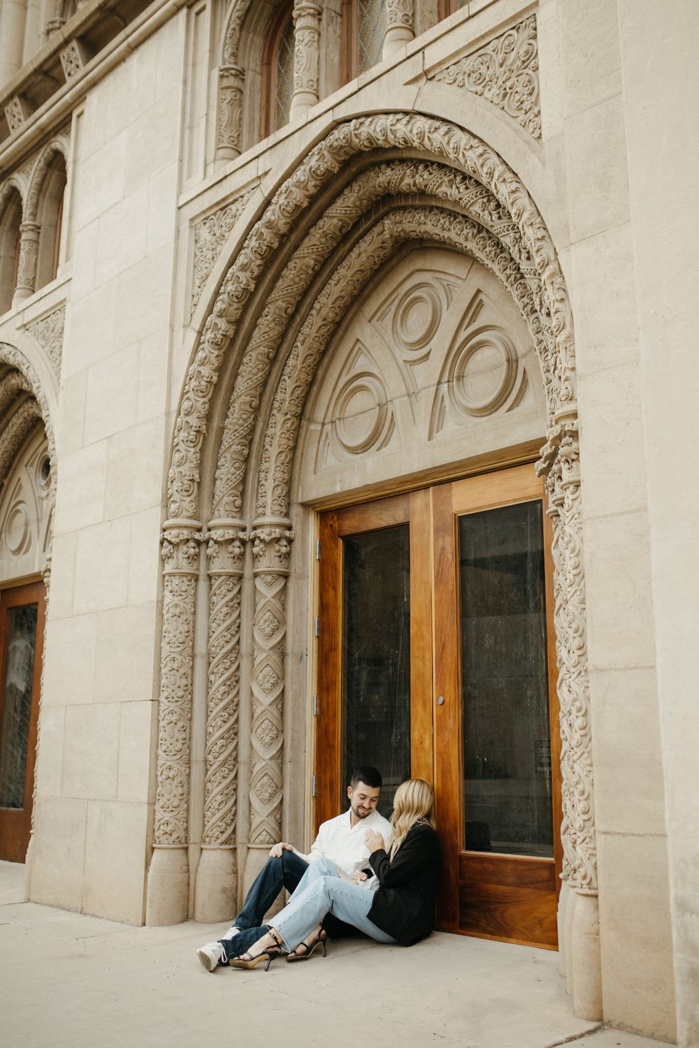 Couple sitting together in front of ornate stone archway with wooden doors at a historic building.