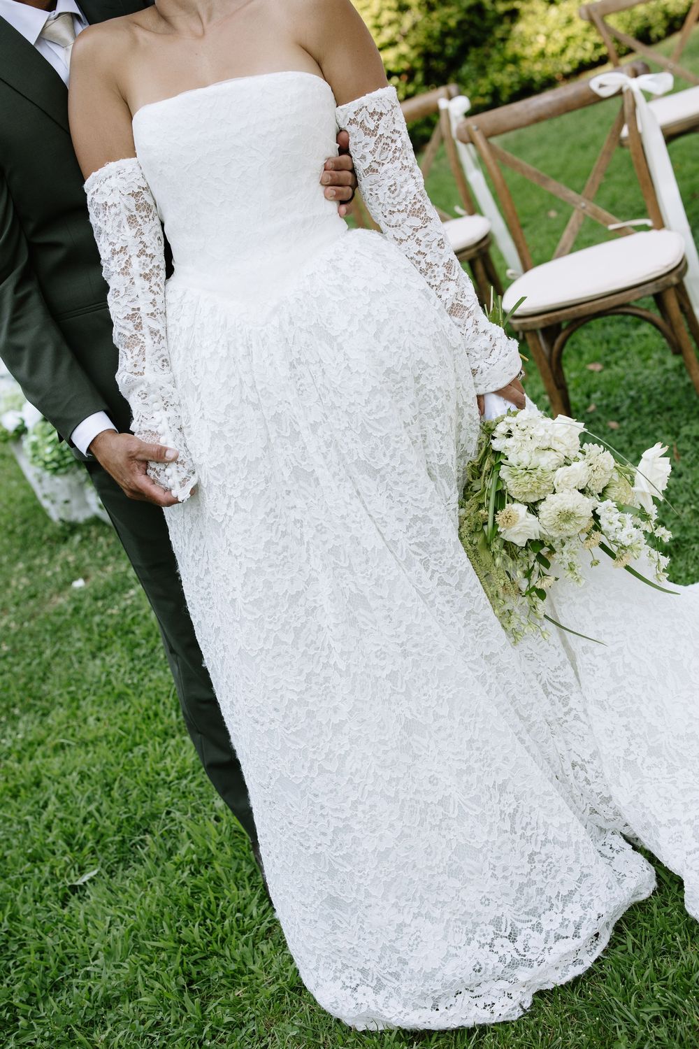 Bride in strapless lace wedding gown with gloves holding white bouquet beside groom in dark suit outdoors.
