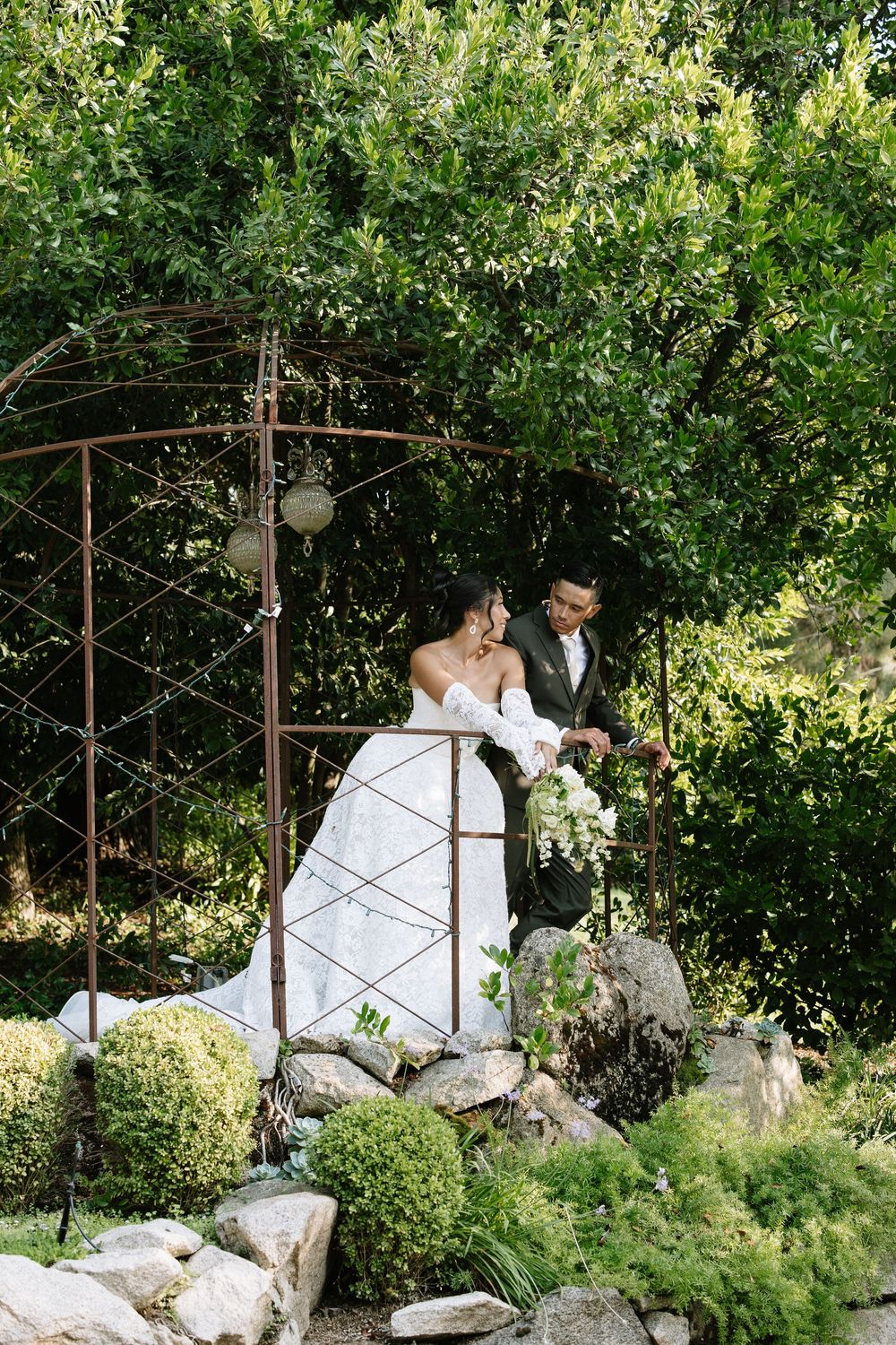 A bride and groom stand under a rustic metal arch surrounded by lush greenery, with the bride holding a white bouquet.
