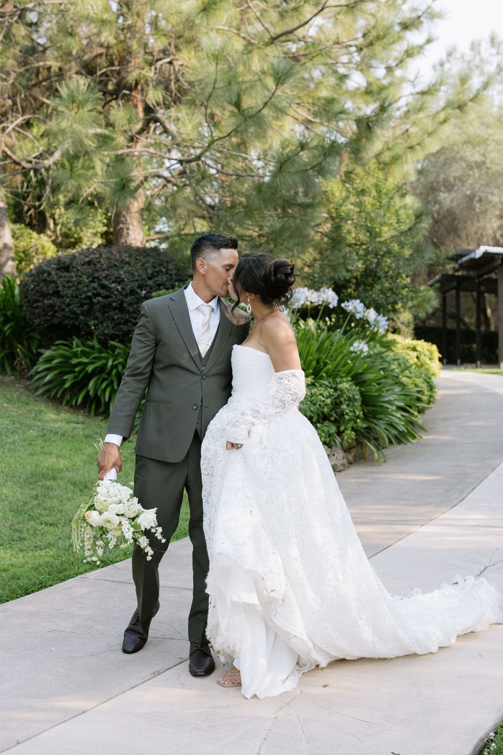 Bride and groom share a kiss on a garden pathway, she in a white ballgown, he in an olive suit holding a bouquet.