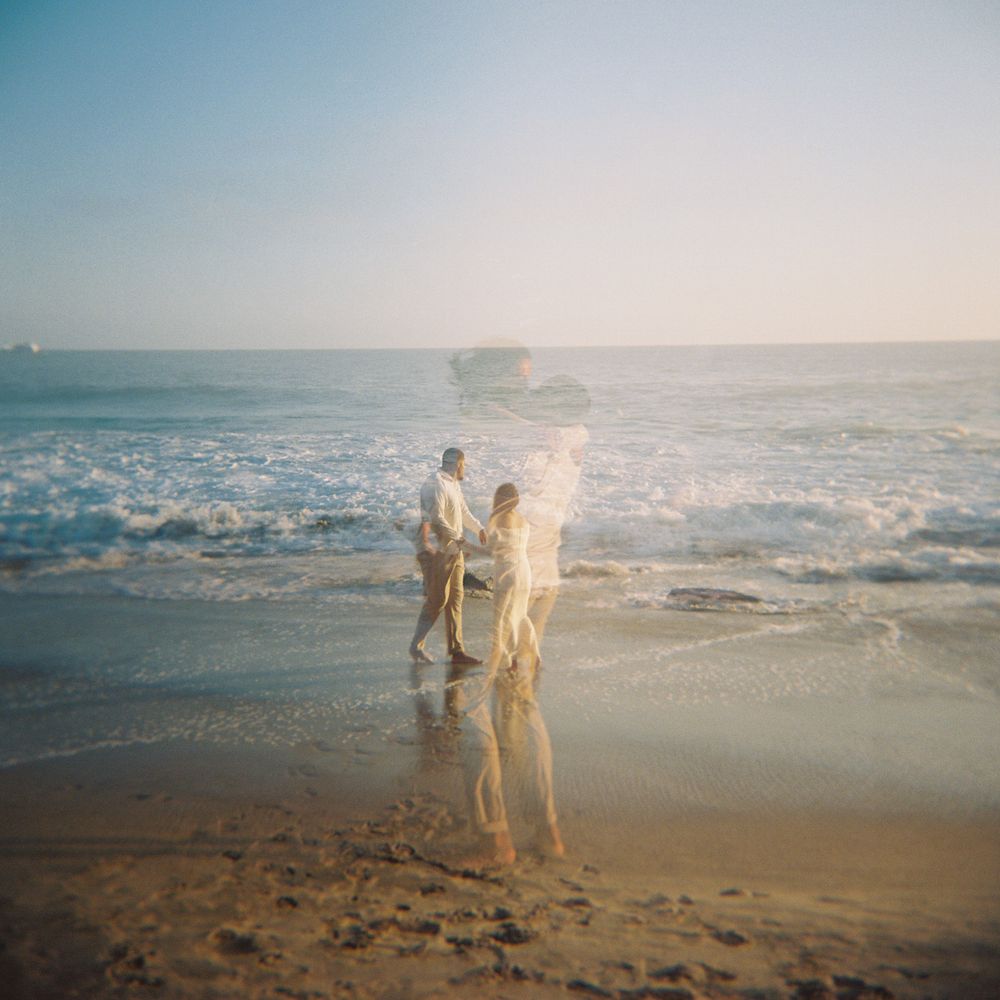 Couple walking hand in hand on a sunlit beach with ocean waves, captured in a dreamy, soft-focus film photography style.