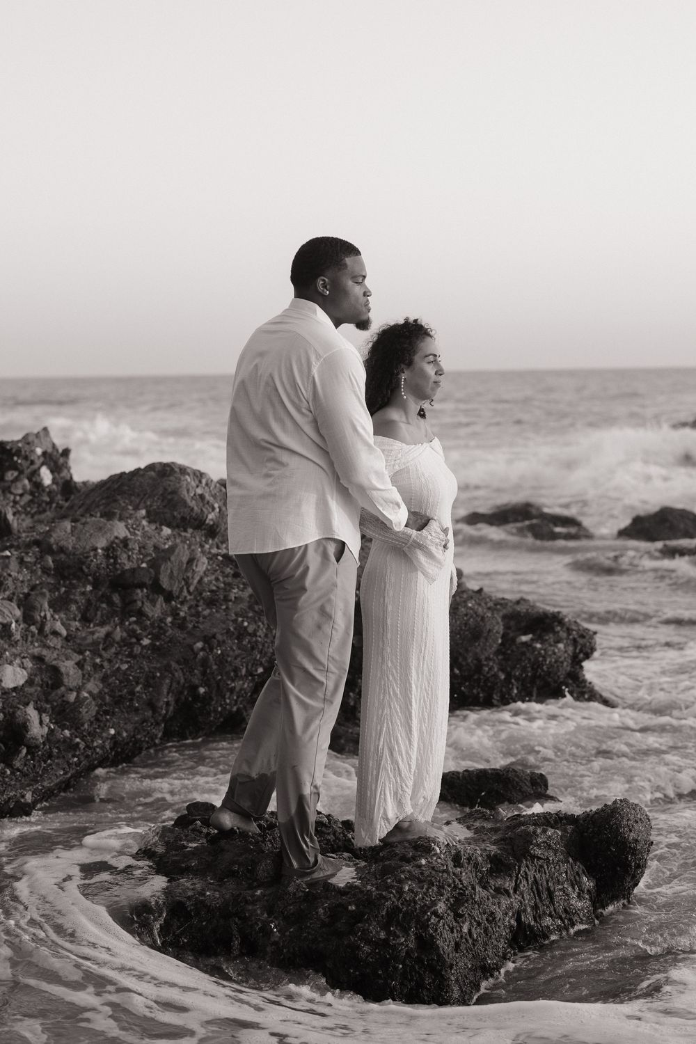Black and white photo of a couple standing on rocks by the ocean, embracing in white outfits.