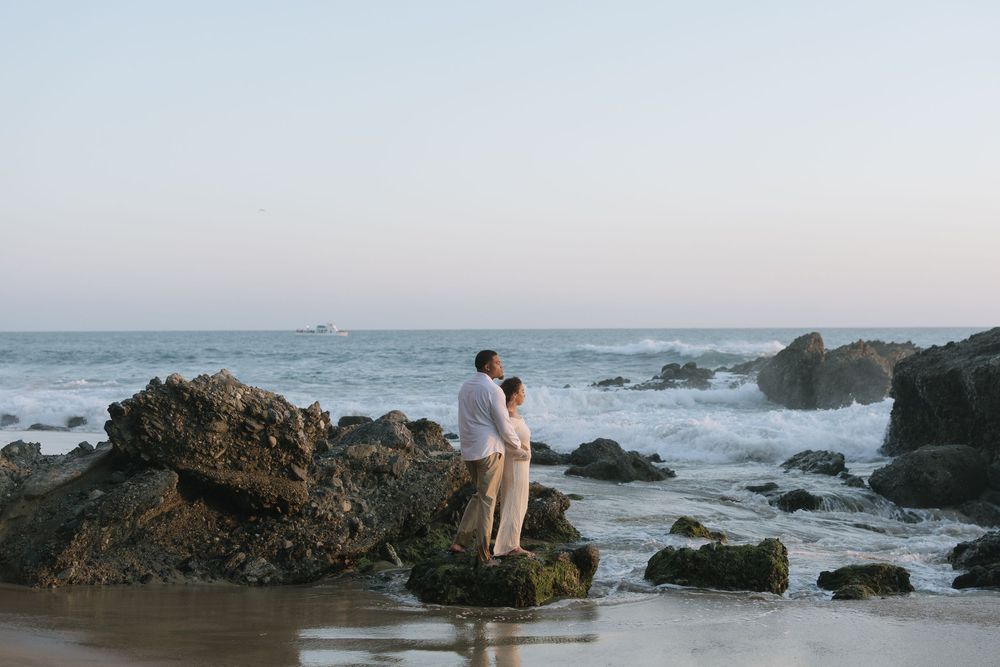 Couple embracing on rocky beach at sunset with ocean waves crashing behind them.
