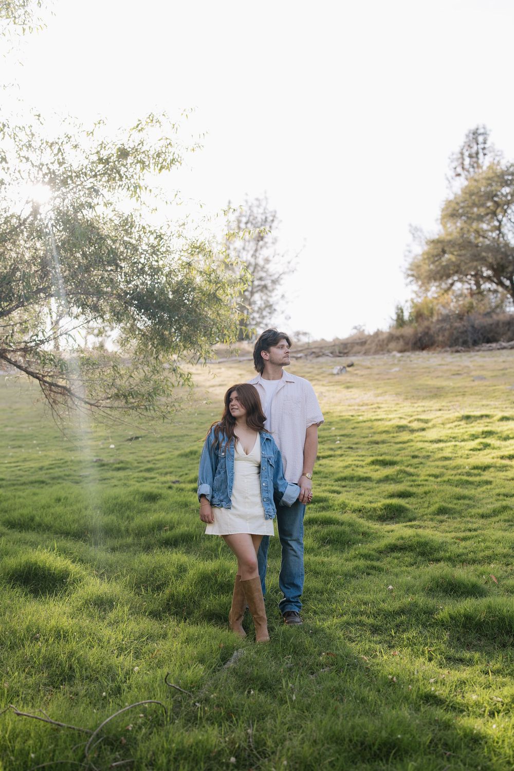 A young couple walks hand in hand on a lush green grassy field surrounded by trees in warm sunlight.