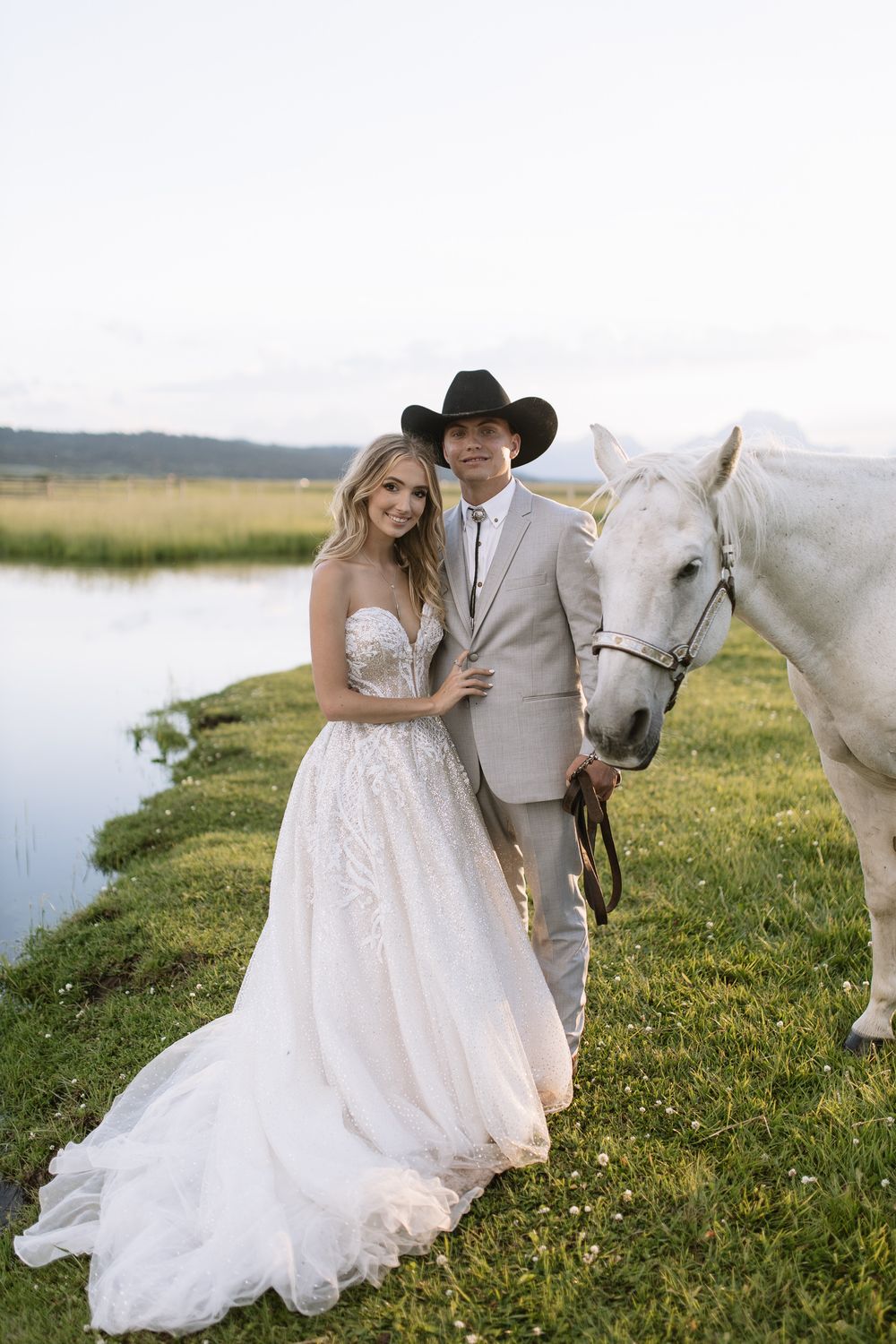 Bride and groom posing with a white horse near a pond, groom wearing a black cowboy hat and gray suit.