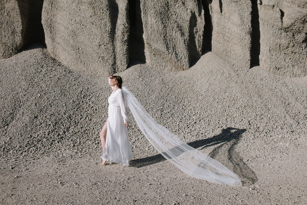 Bride in white gown with long flowing veil stands among dramatic grey sand dunes in an ethereal outdoor wedding shoot.