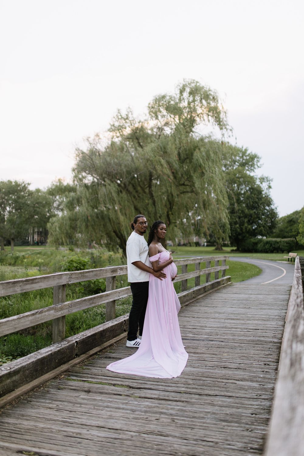Couple poses on wooden bridge, pregnant woman in flowing pink gown, surrounded by lush greenery and willow trees in Sacramento, California.