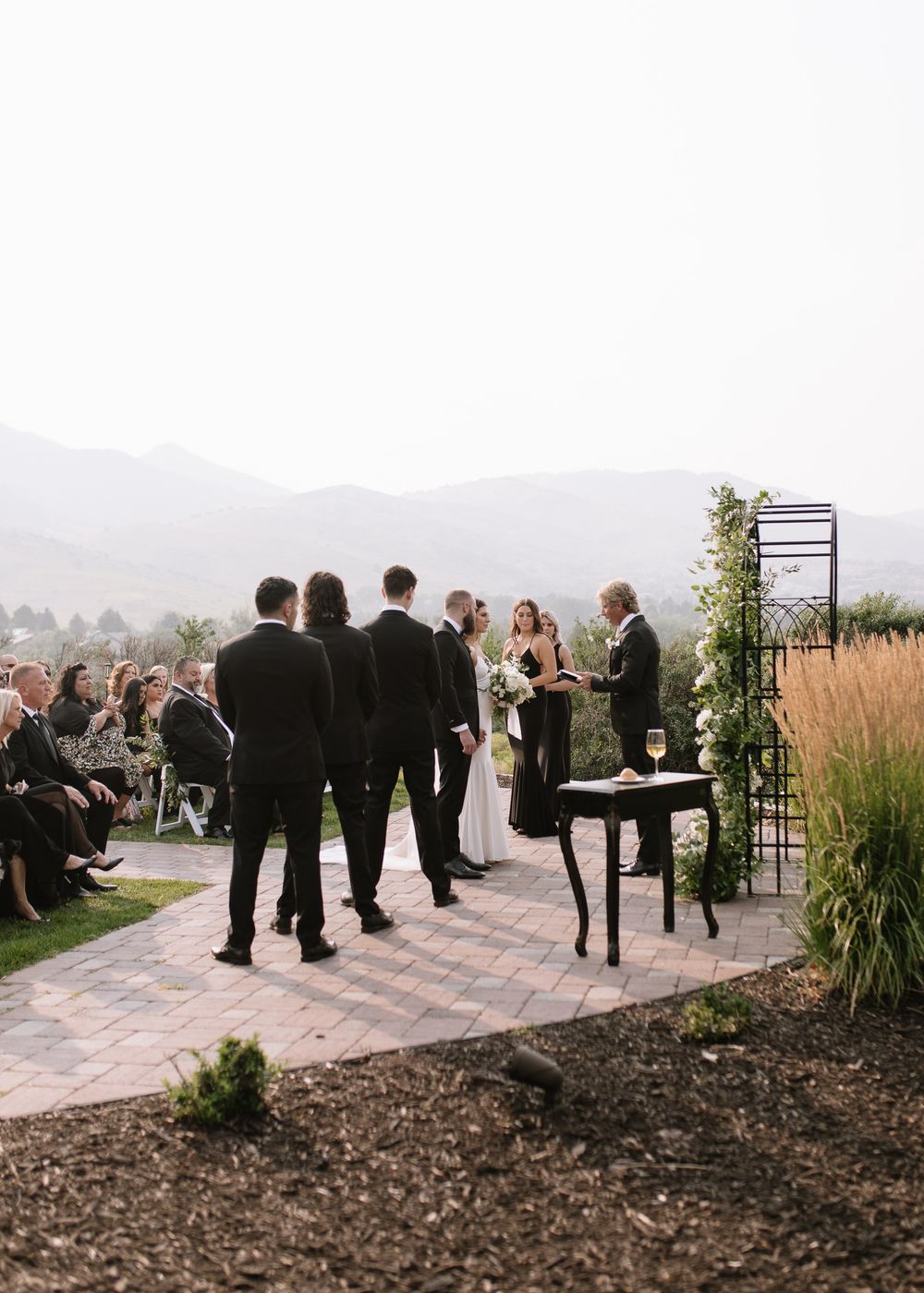 Outdoor wedding ceremony on a brick patio with mountain views, guests seated, bridal party in black suits near floral arch.