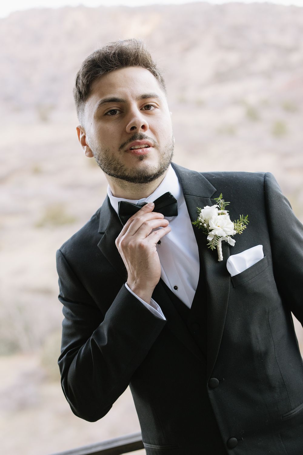 A groom in a black tuxedo adjusts his bow tie, wearing a white boutonniere, with a desert landscape backdrop in Northern California.