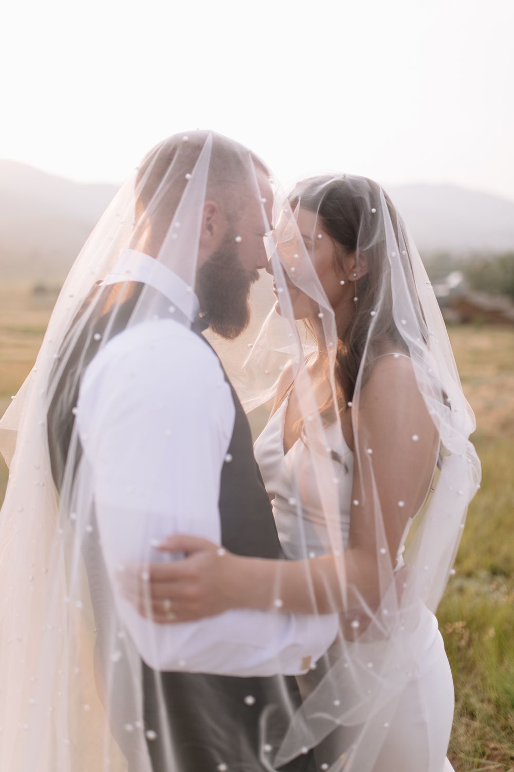 A bride and groom share a romantic kiss under a pearl-adorned wedding veil in a scenic outdoor setting.
