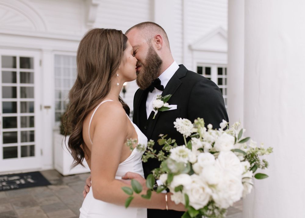 A bride and groom share a romantic kiss outside a white venue, with the bride holding a lush white floral bouquet.