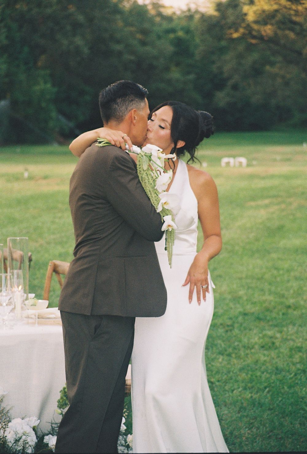 Bride and groom share a romantic kiss outdoors at their wedding reception, surrounded by lush greenery.