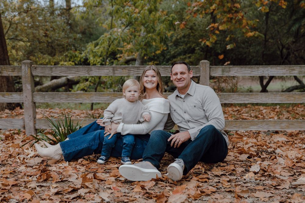 A family sits together on fallen autumn leaves in a park setting during a casual outdoor photo session.