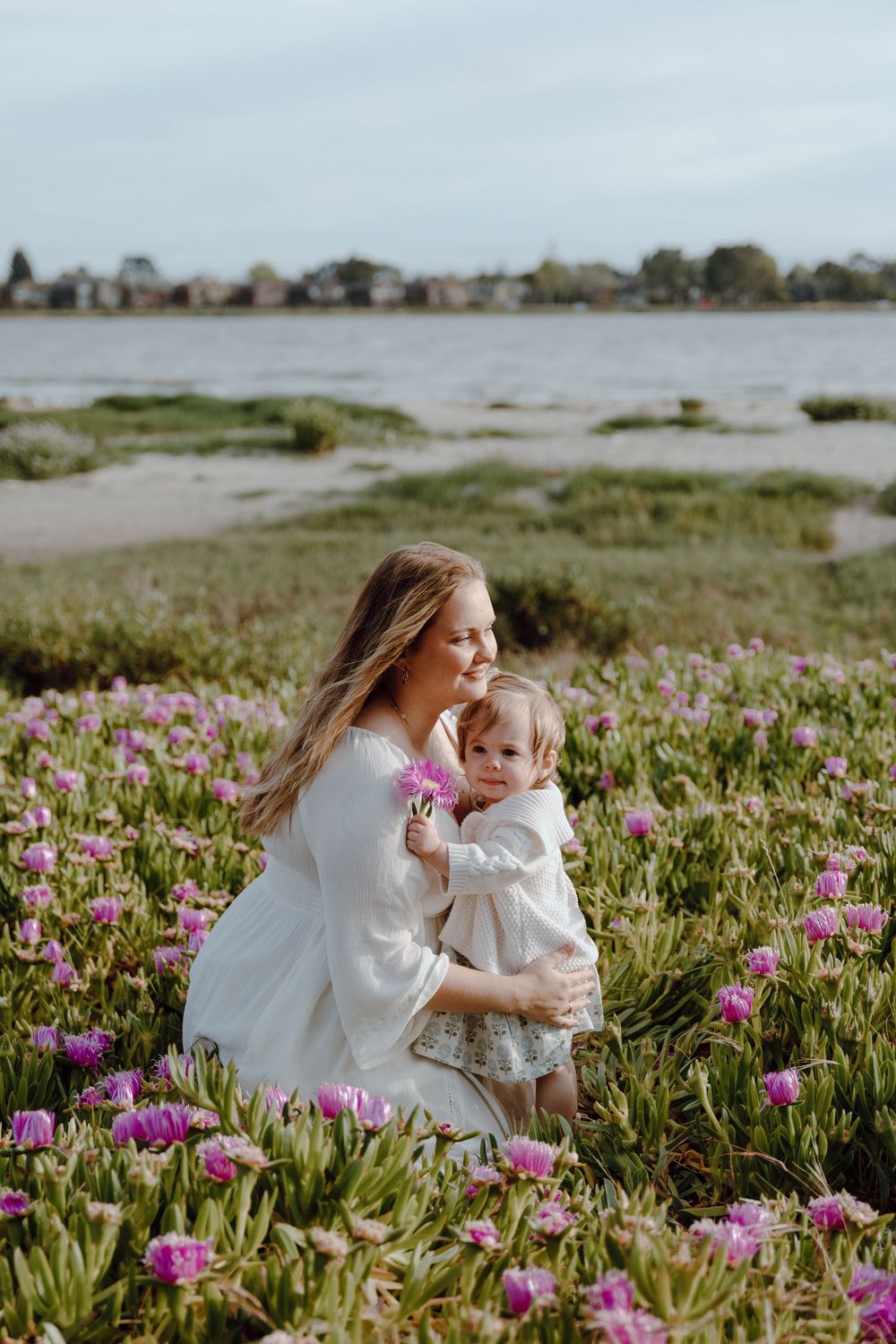 Two people in white dresses sit among vibrant purple wildflowers blooming in a coastal meadow near the ocean.
