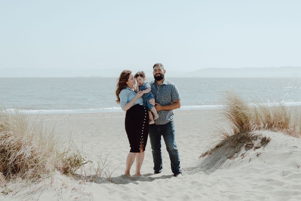 A family embraces while standing on sand dunes near the ocean on a bright day.