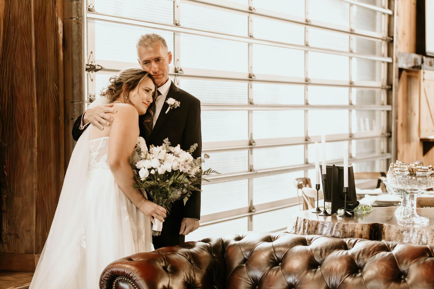 A romantic wedding reception setup with rustic wooden beams and soft natural lighting through large windows.