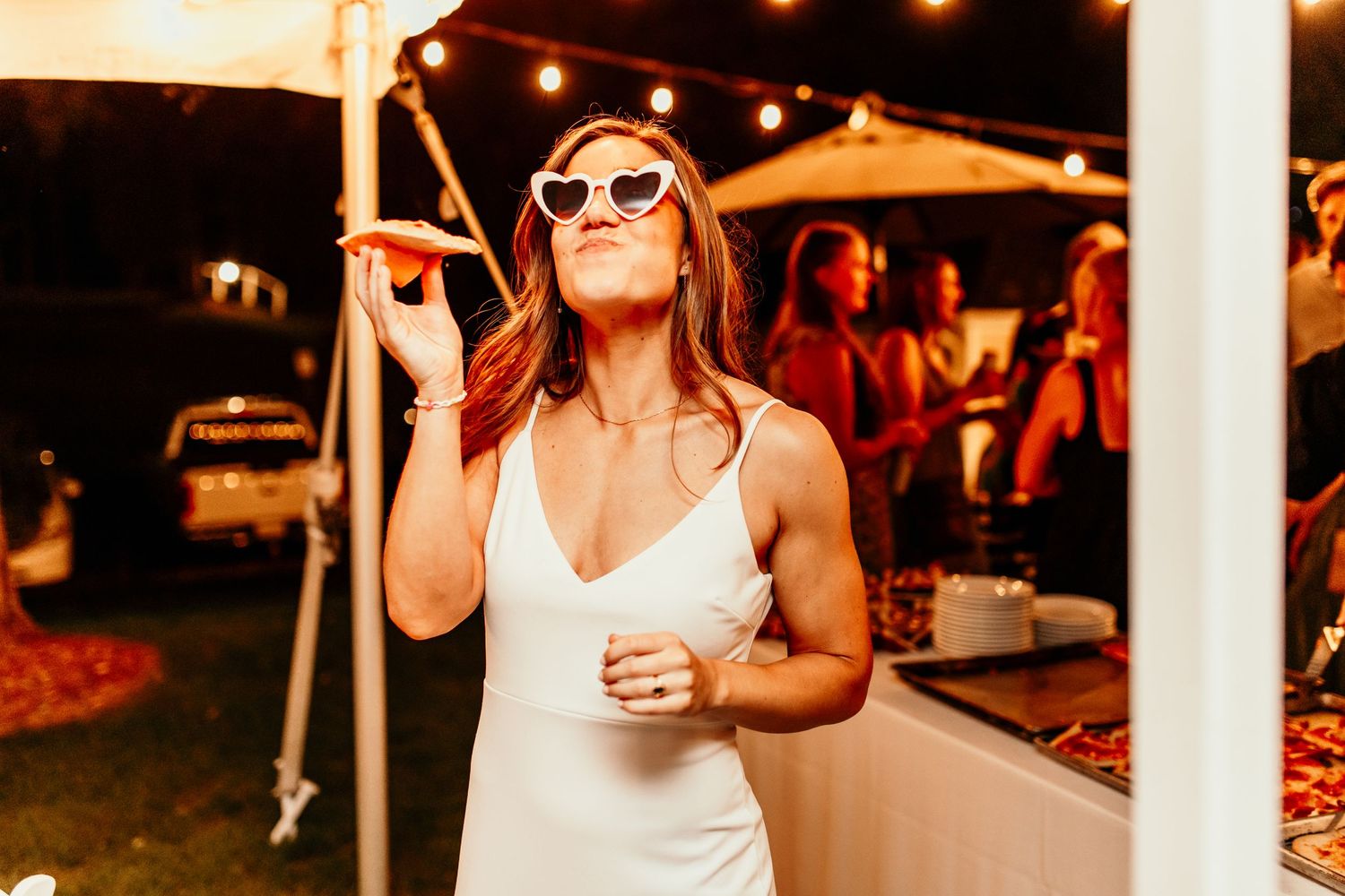 Woman in white dress dances with sunglasses at outdoor evening reception.