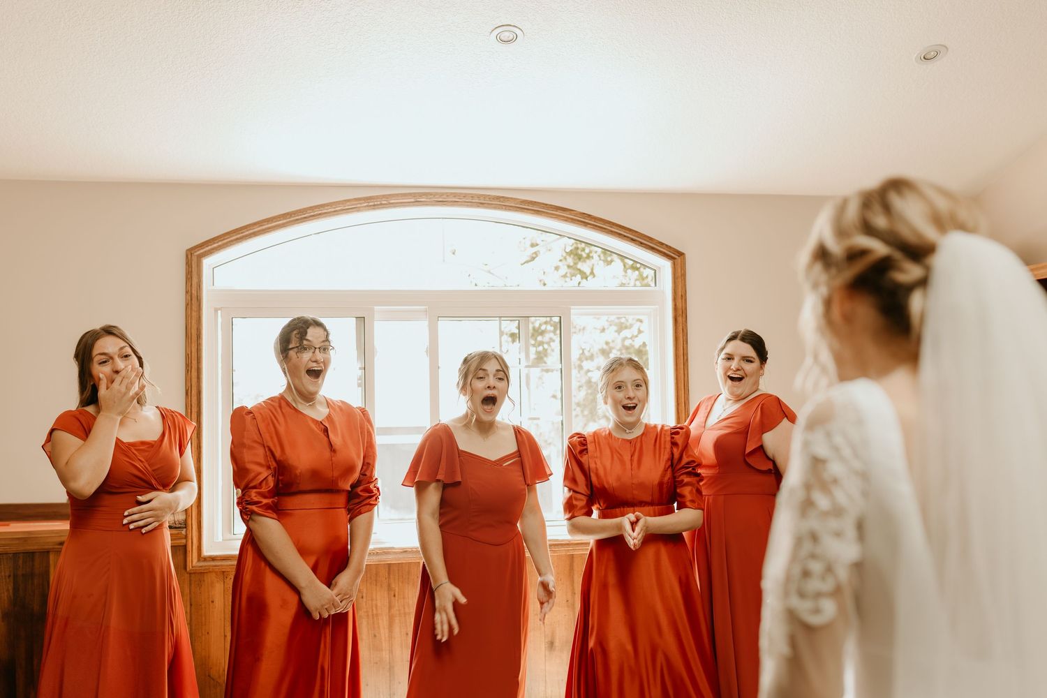 A group wearing matching red dresses stands together in a bridal preparation room with natural lighting.