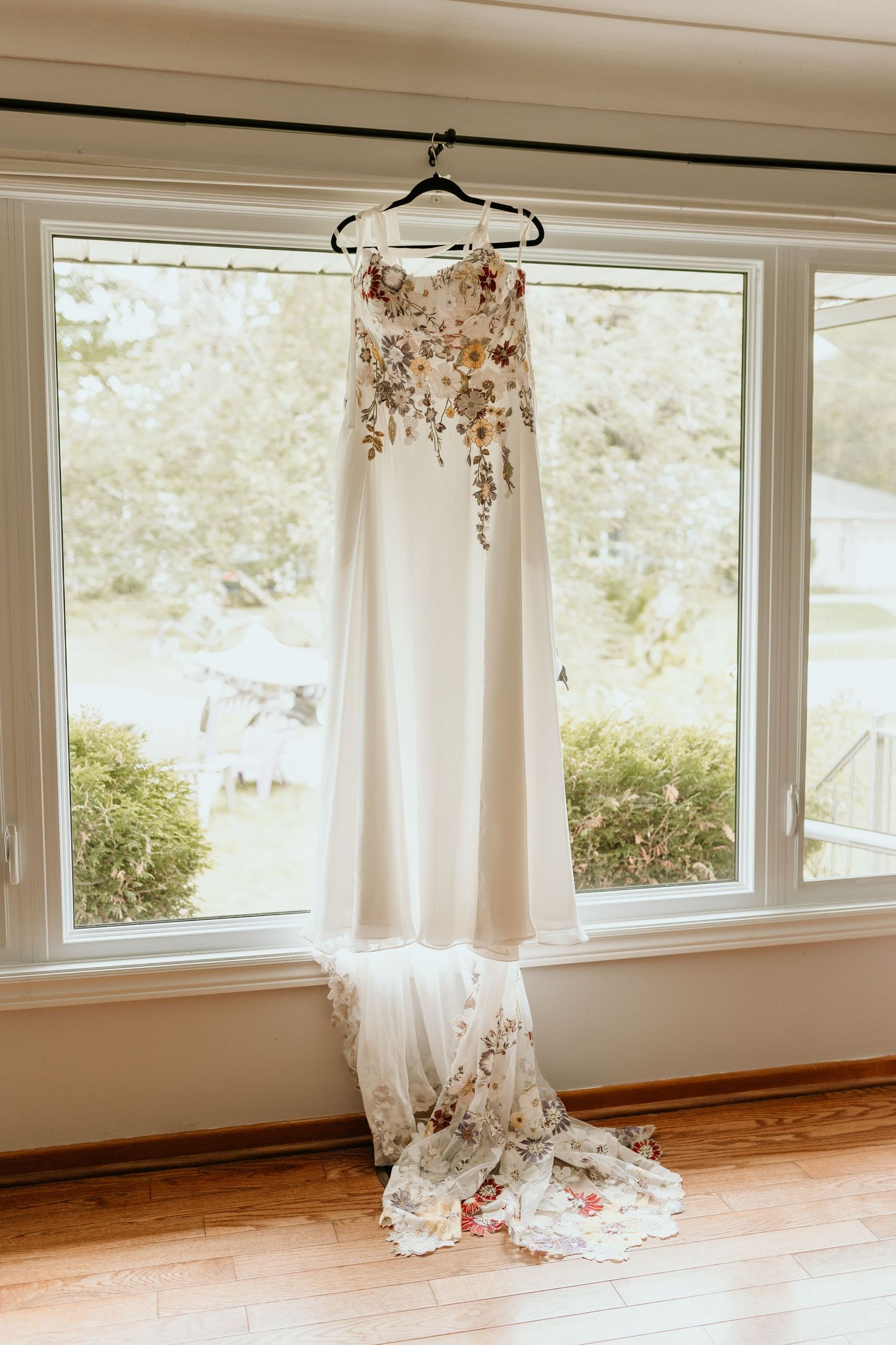White wedding dress hangs in a sunlit room with large windows and hardwood floors, creating an ethereal bridal scene.