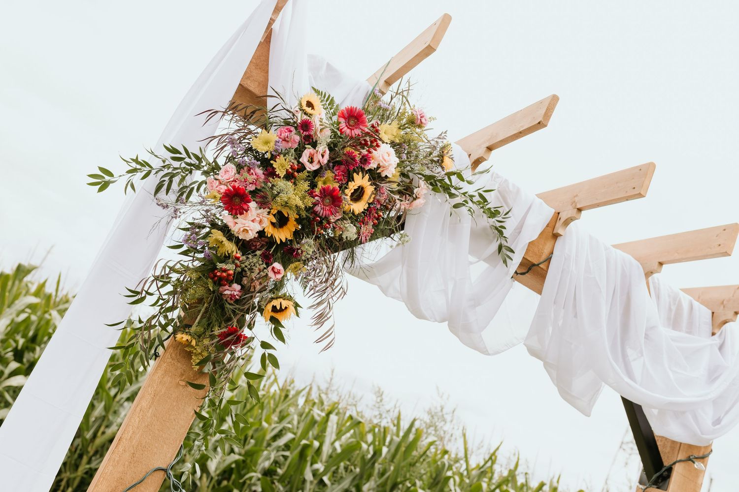 A wooden wedding arch decorated with pink and peach flowers and white fabric draping stands in a grassy field.