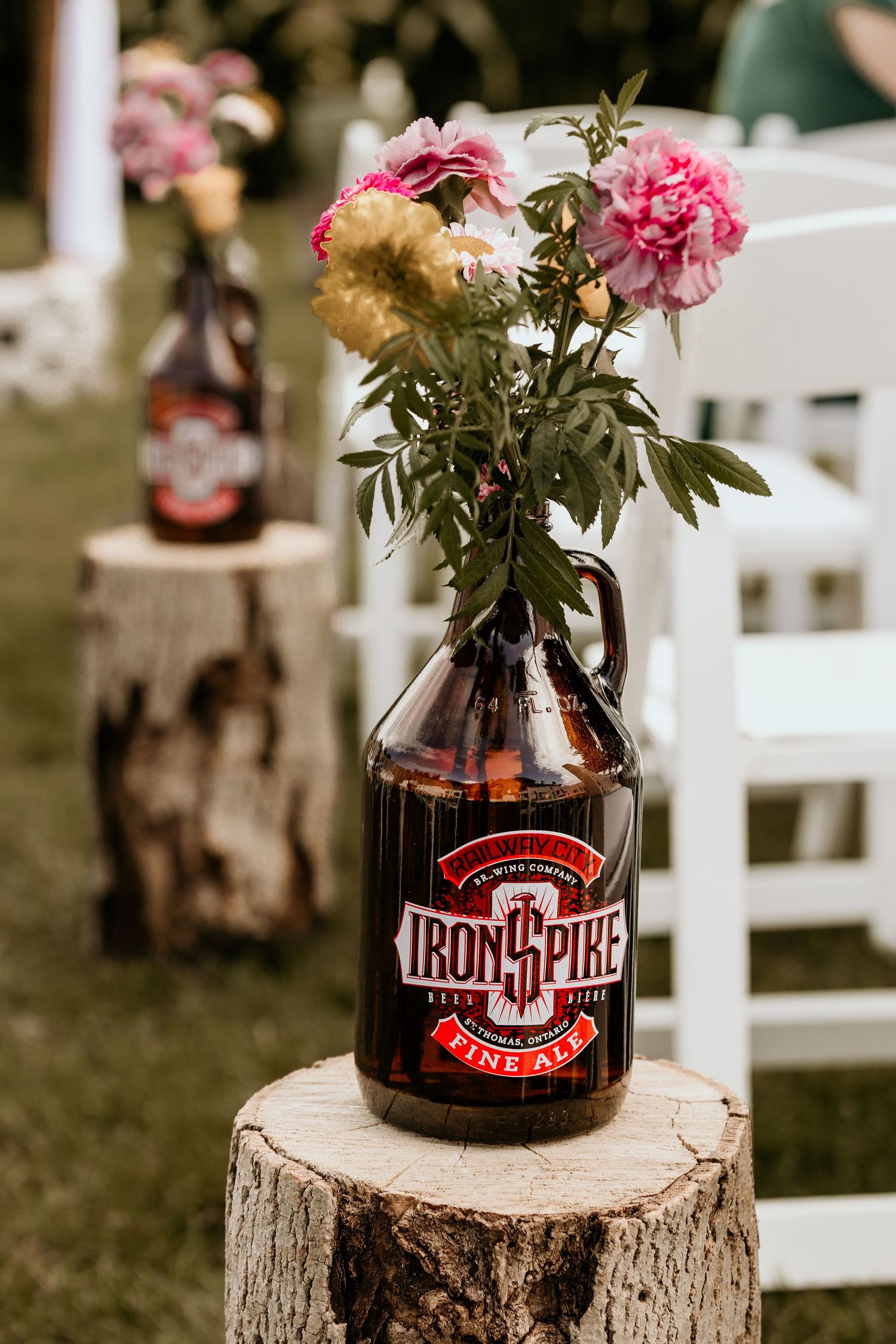 Pink flowers arranged in repurposed Iron Horse beer bottles displayed on rustic wood stumps at an outdoor event setting.