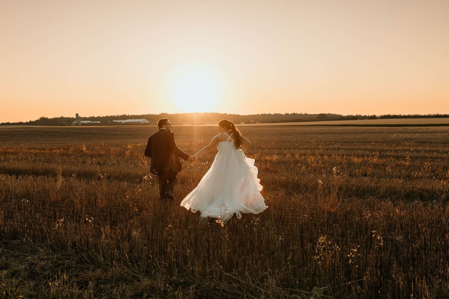 Two silhouettes walk through a golden field at sunset with a glowing white dress flowing in the evening light.