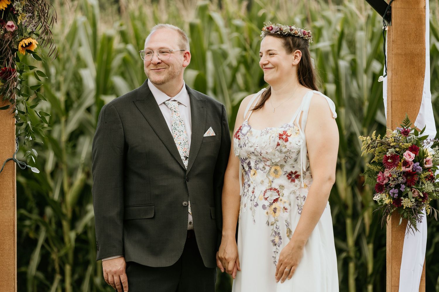 A couple shares a joyful moment at their outdoor wedding ceremony with a cornfield backdrop and floral decorations.