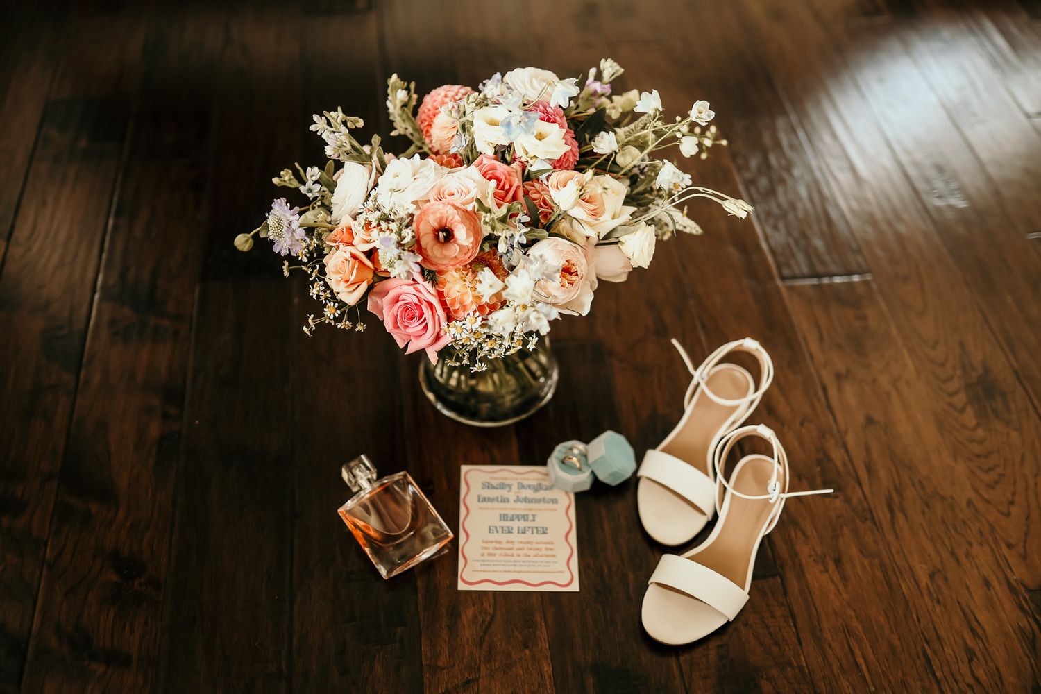 Wedding details flat lay with peach floral arrangement, bridal shoes and invitation on wooden floor.