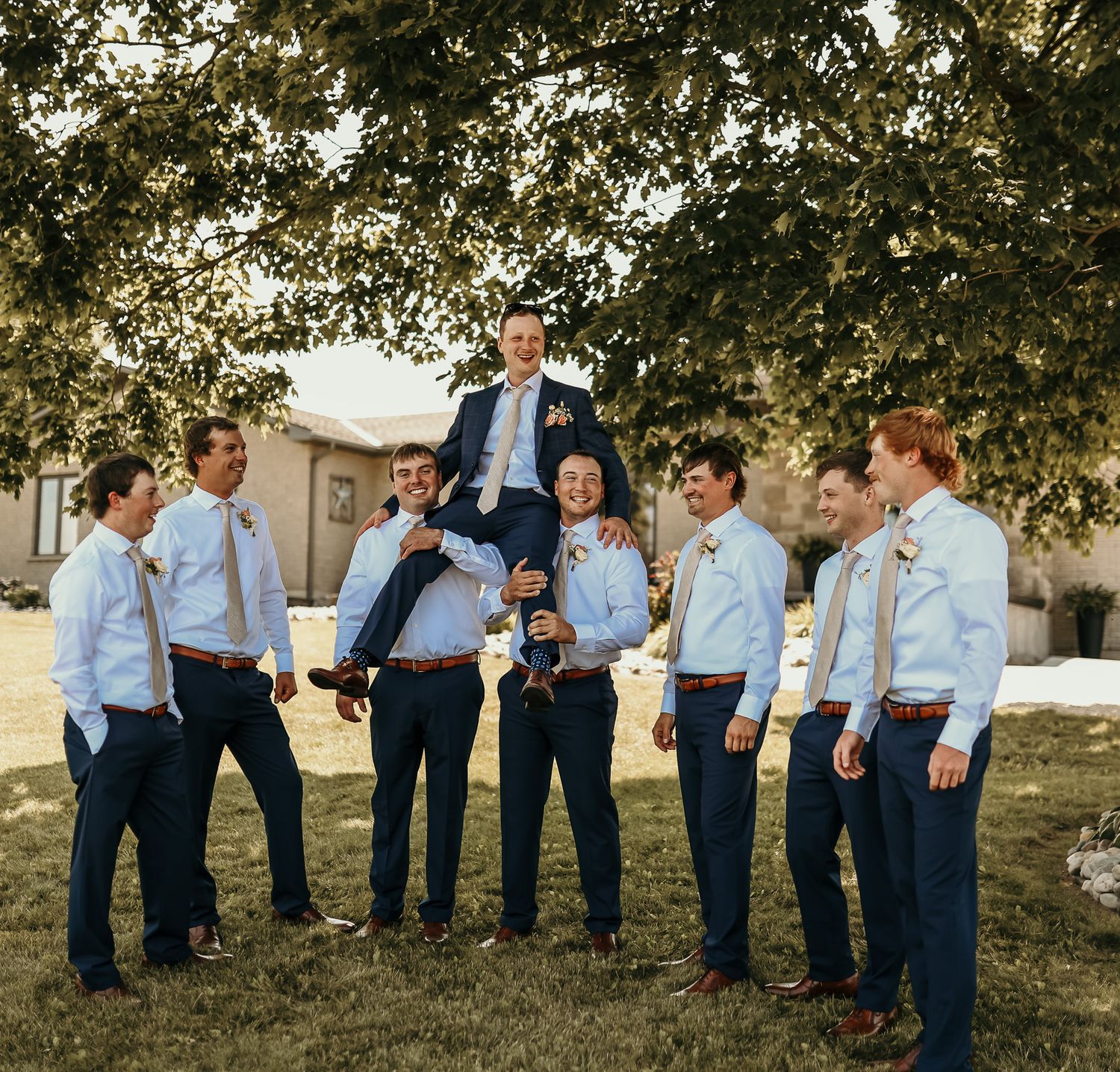 Groomsmen in light blue shirts lifting the groom during an outdoor wedding photo.