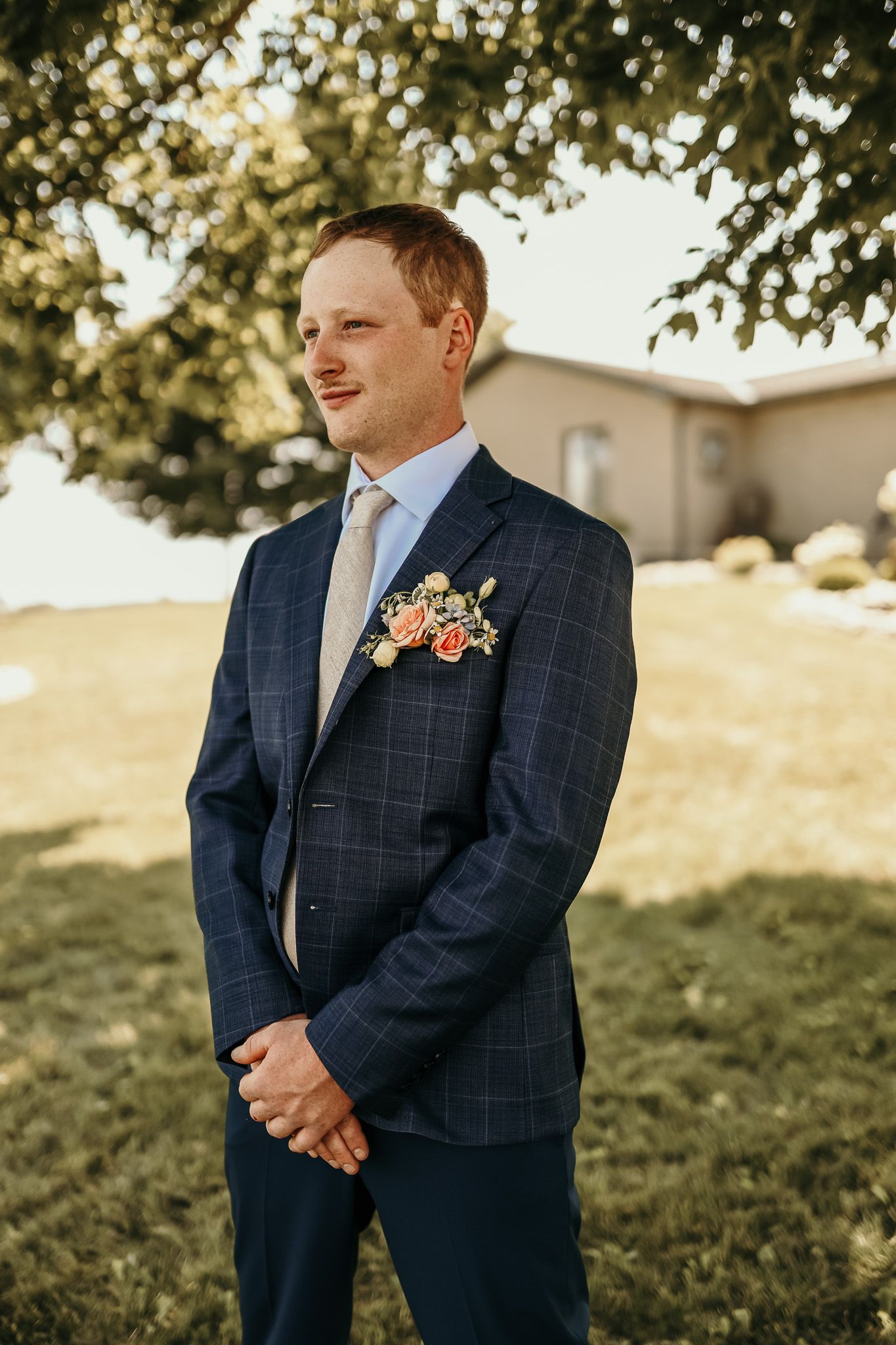 Wedding attire featuring navy checkered suit jacket with floral boutonniere against outdoor garden backdrop.