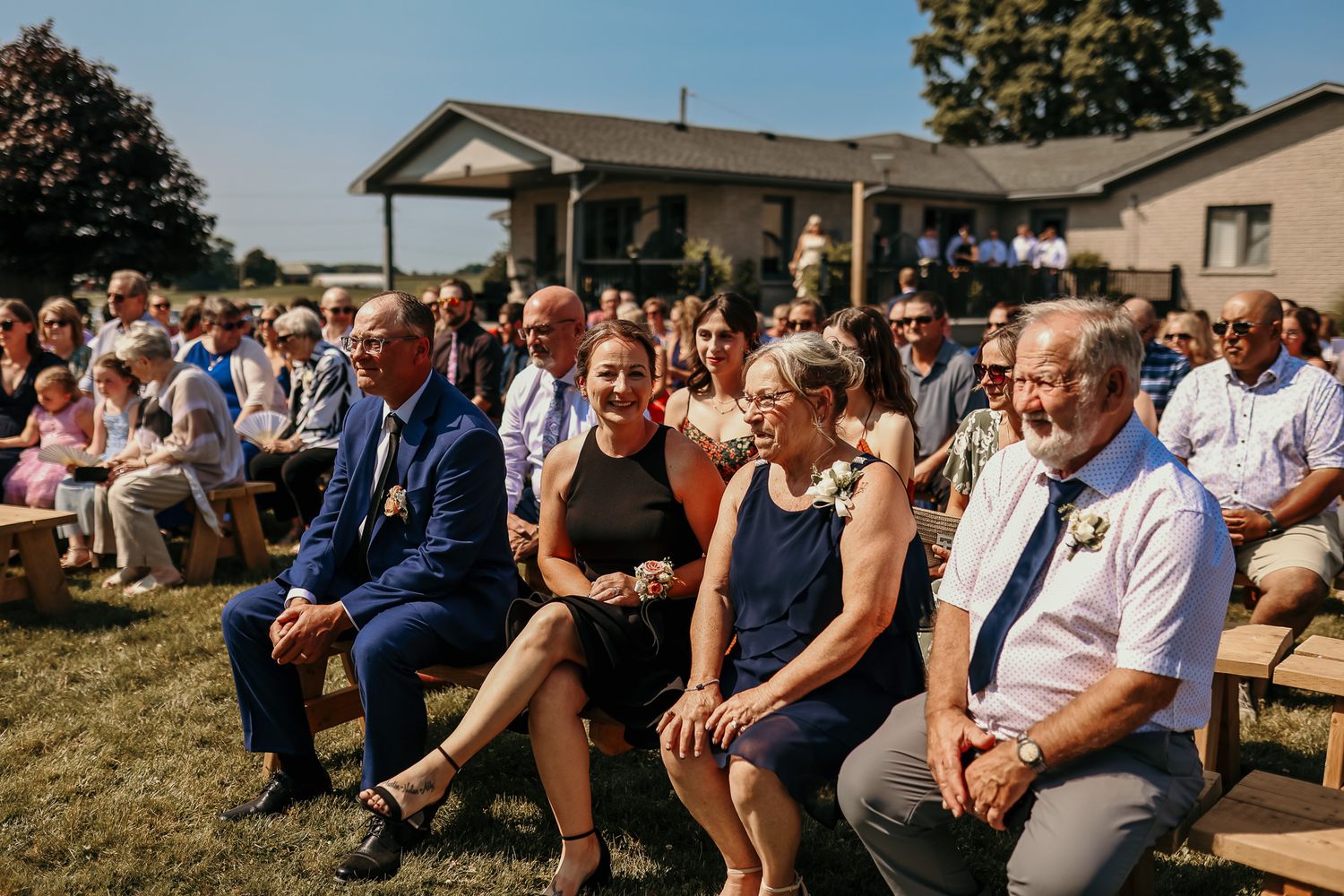 Outdoor wedding ceremony guests seated on wooden chairs under blue summer sky.