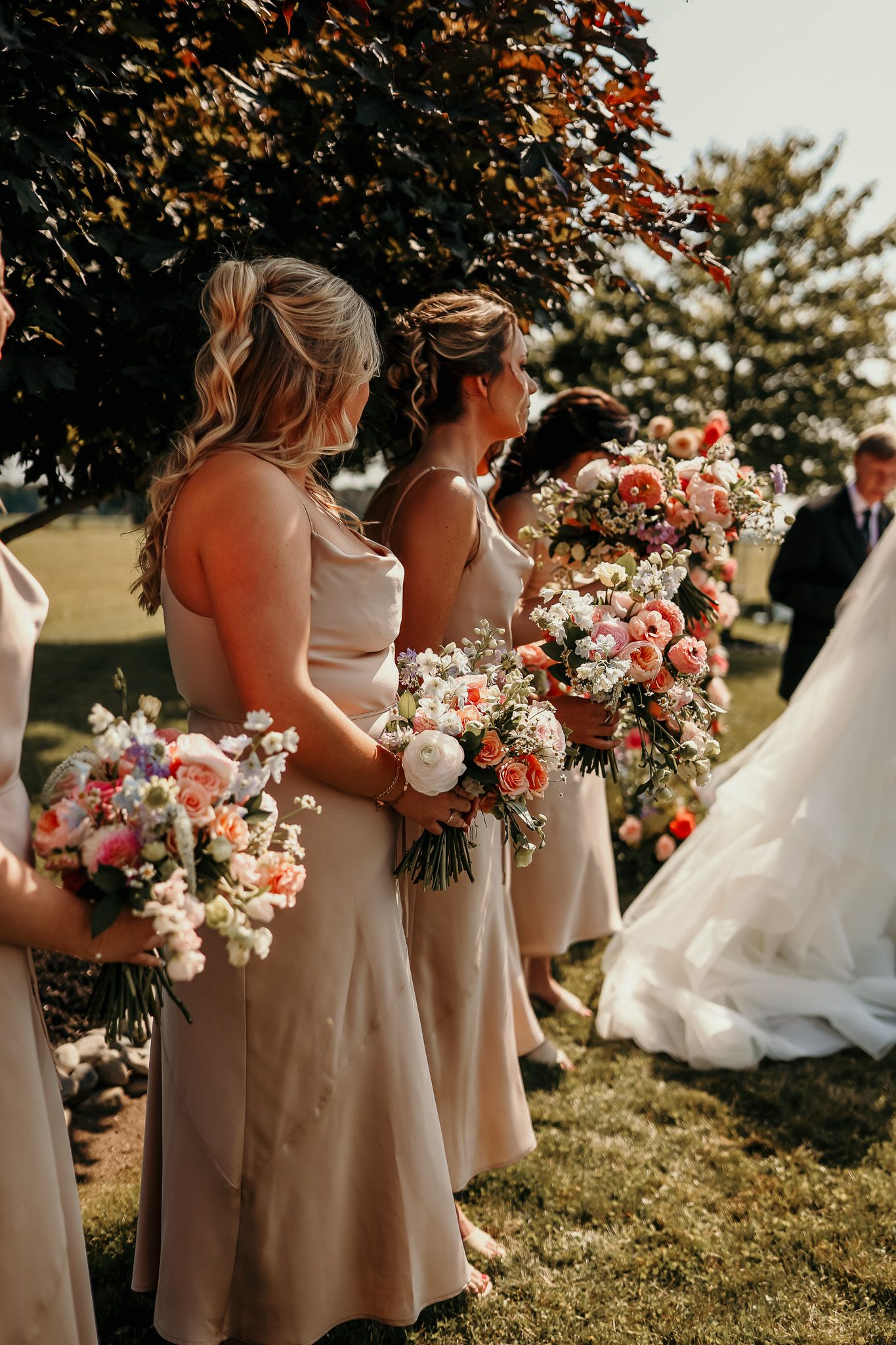 Bridesmaids in blush dresses holding pink and white bouquets during outdoor ceremony.