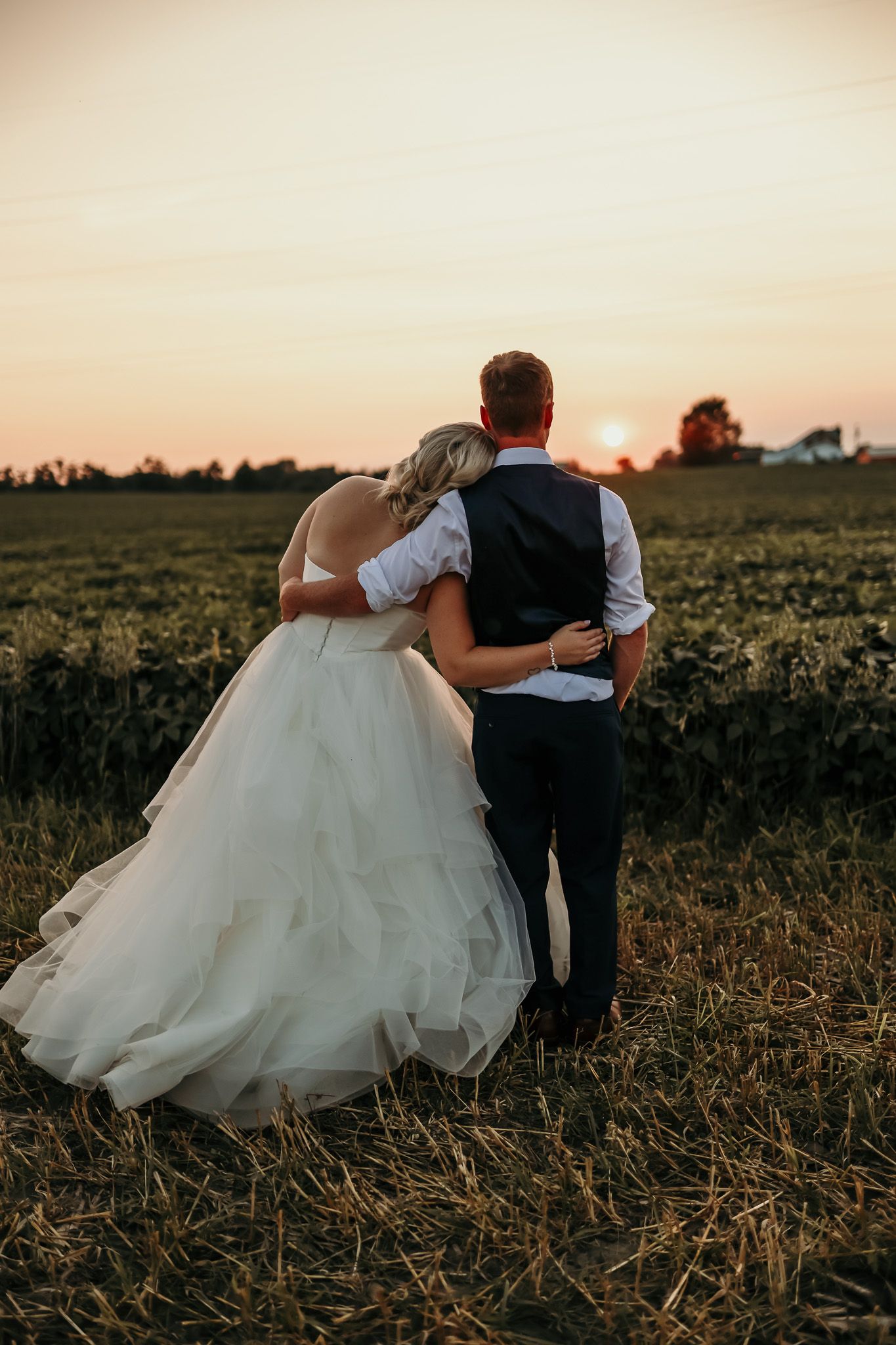 Silhouetted newlyweds embracing while watching sunset in rural field.