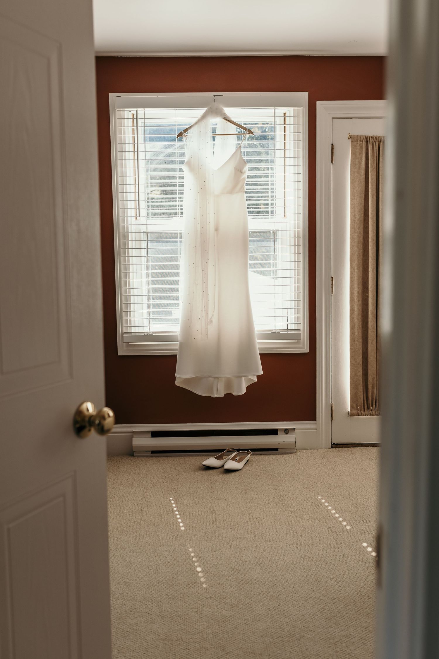 Elegant white wedding dress hanging in window light with shoes on carpet below.