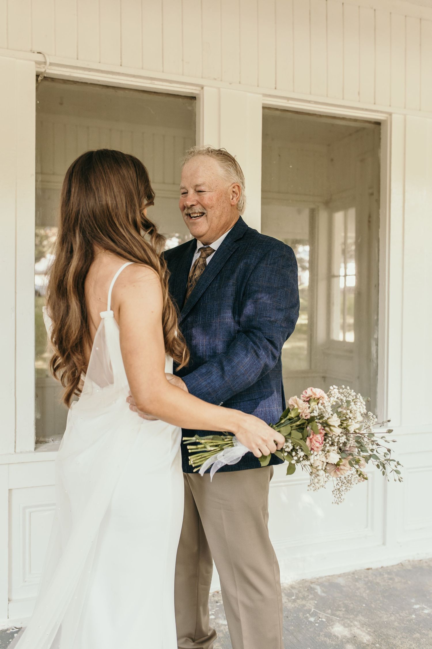 Sweet moment between a bride in white dress and older man with bouquet on a porch.