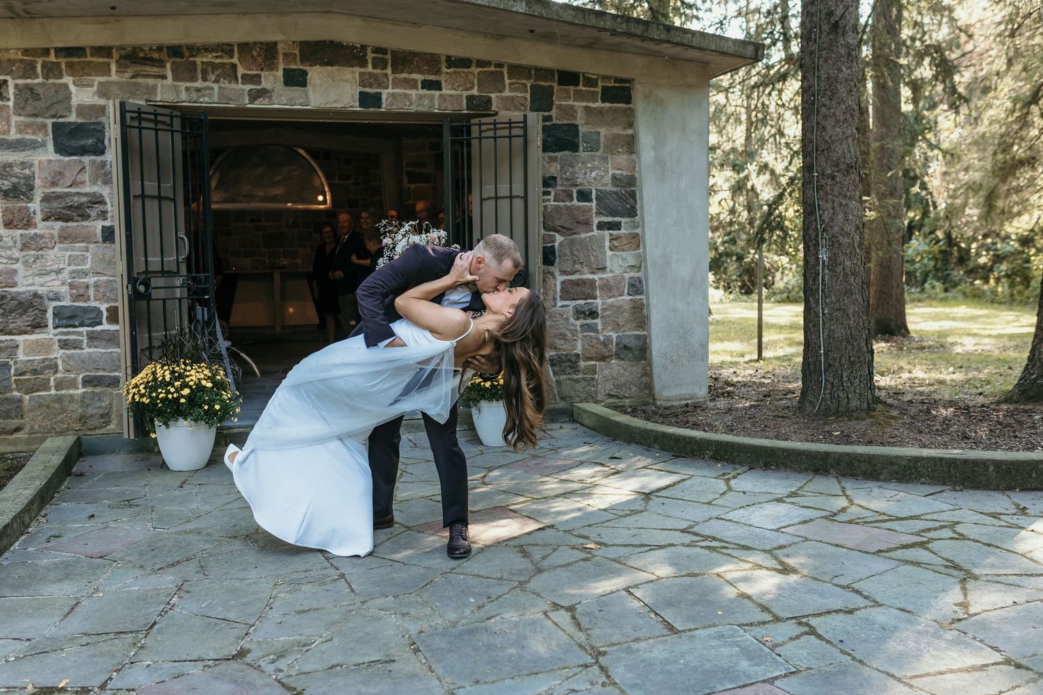 Couple performs romantic dip pose outside stone building during wedding photoshoot.