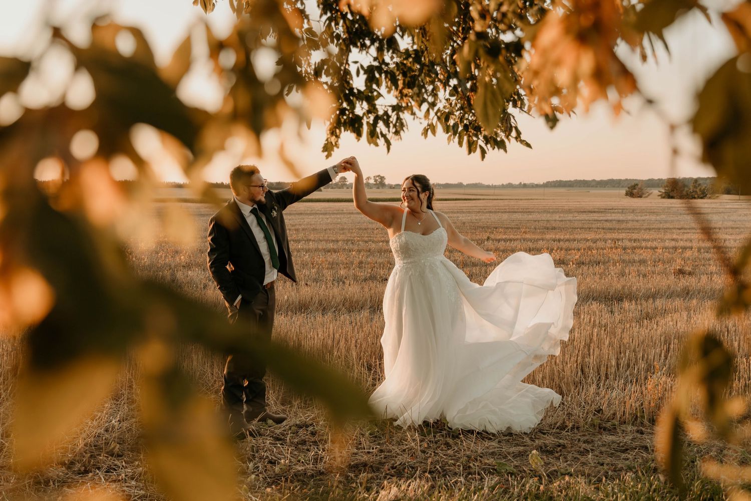A couple in wedding attire dances in a golden wheat field at sunset creating a romantic silhouette.