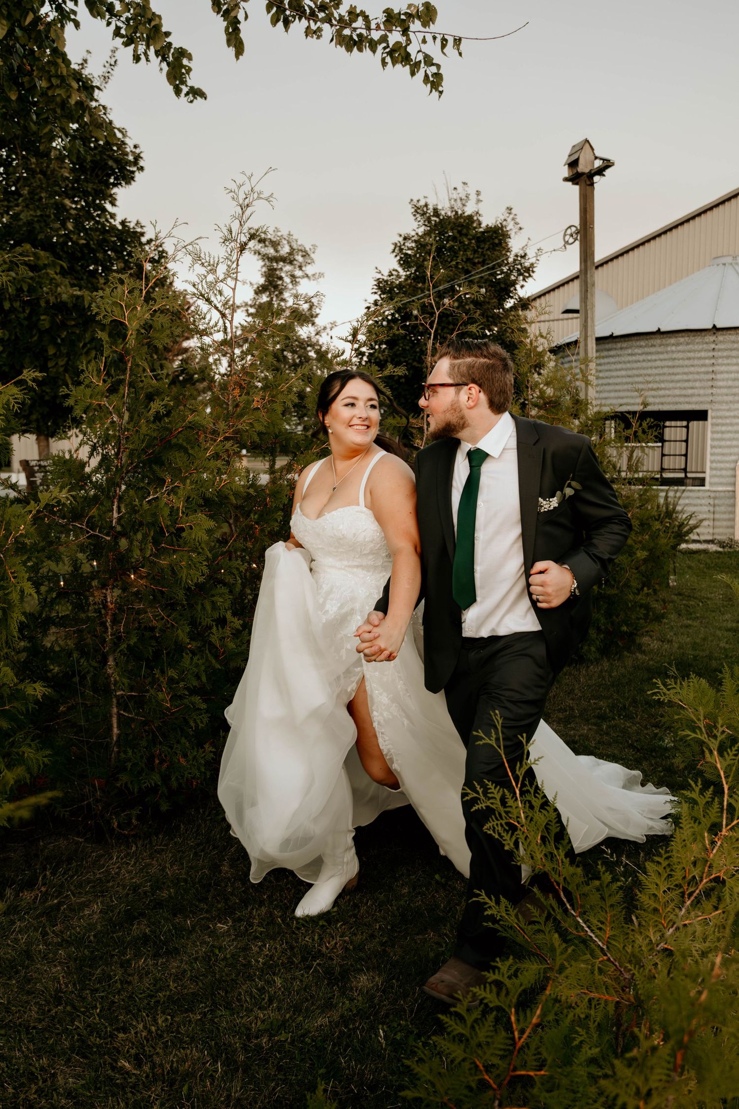 A couple in wedding attire run and laugh together through a garden on their special day.