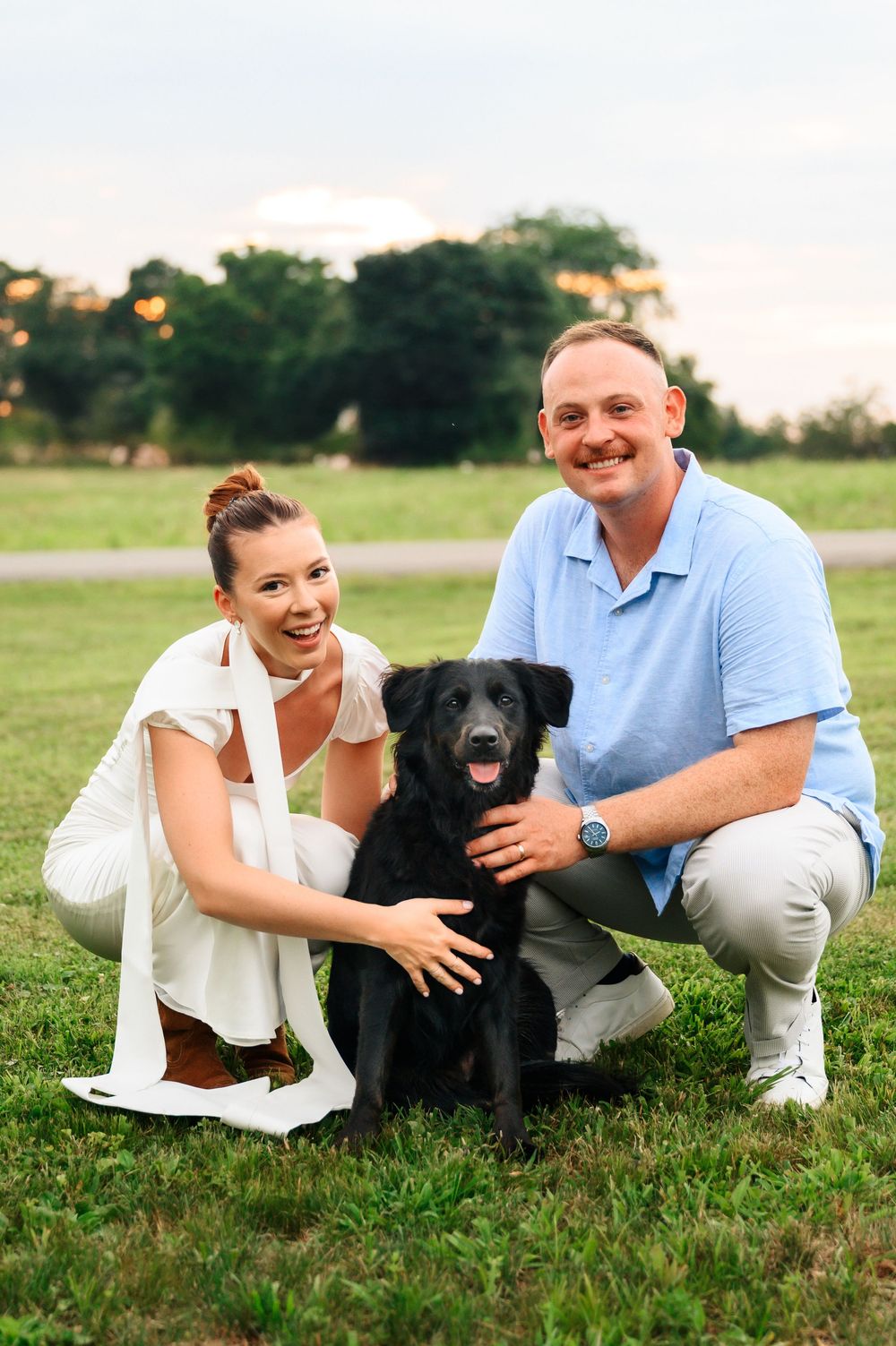 A happy couple poses outdoors on grass with their smiling black dog during a sunny evening.