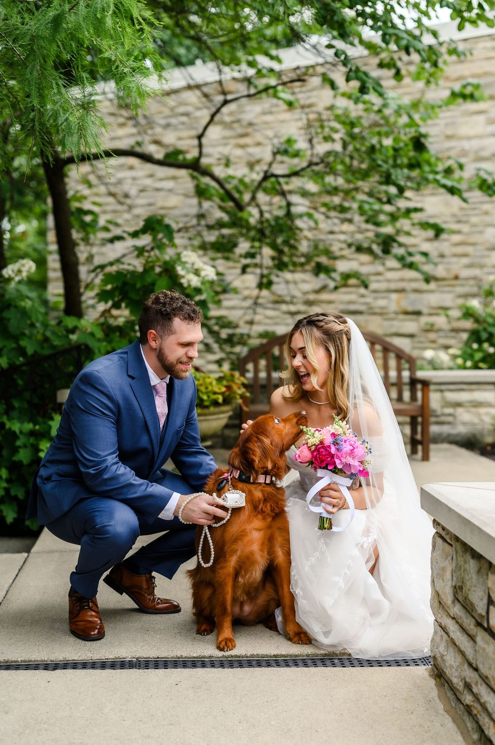 Bride and groom in wedding attire crouching with their golden Irish Setter dog outdoors near stone wall.