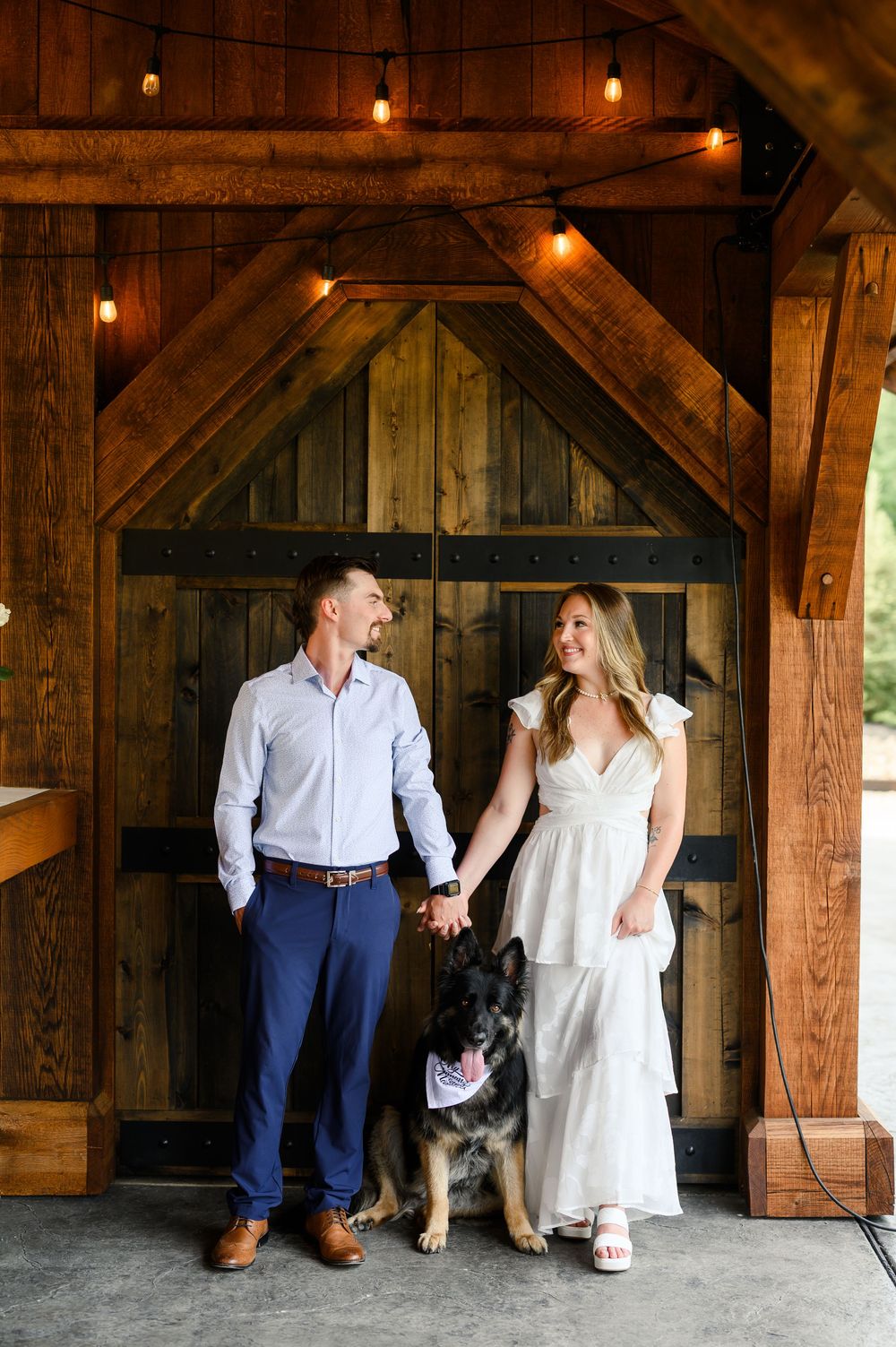 Couple holding hands with German Shepherd dog in front of rustic wooden barn doors with string lights above.