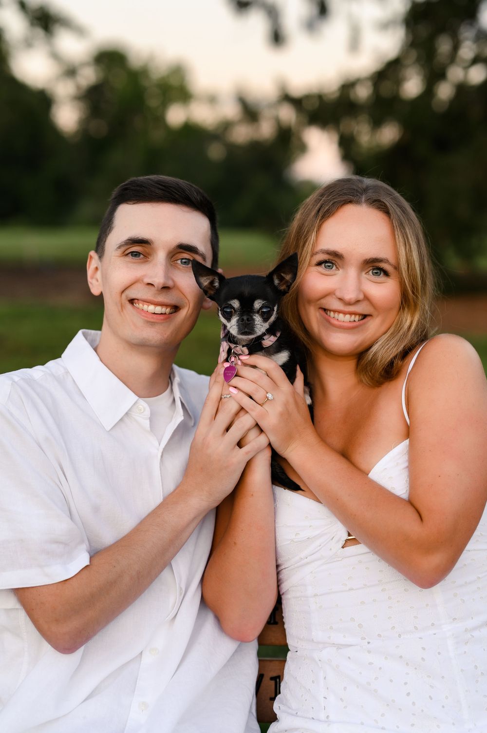 A smiling couple in white outfits holding a small black Chihuahua outdoors at sunset.