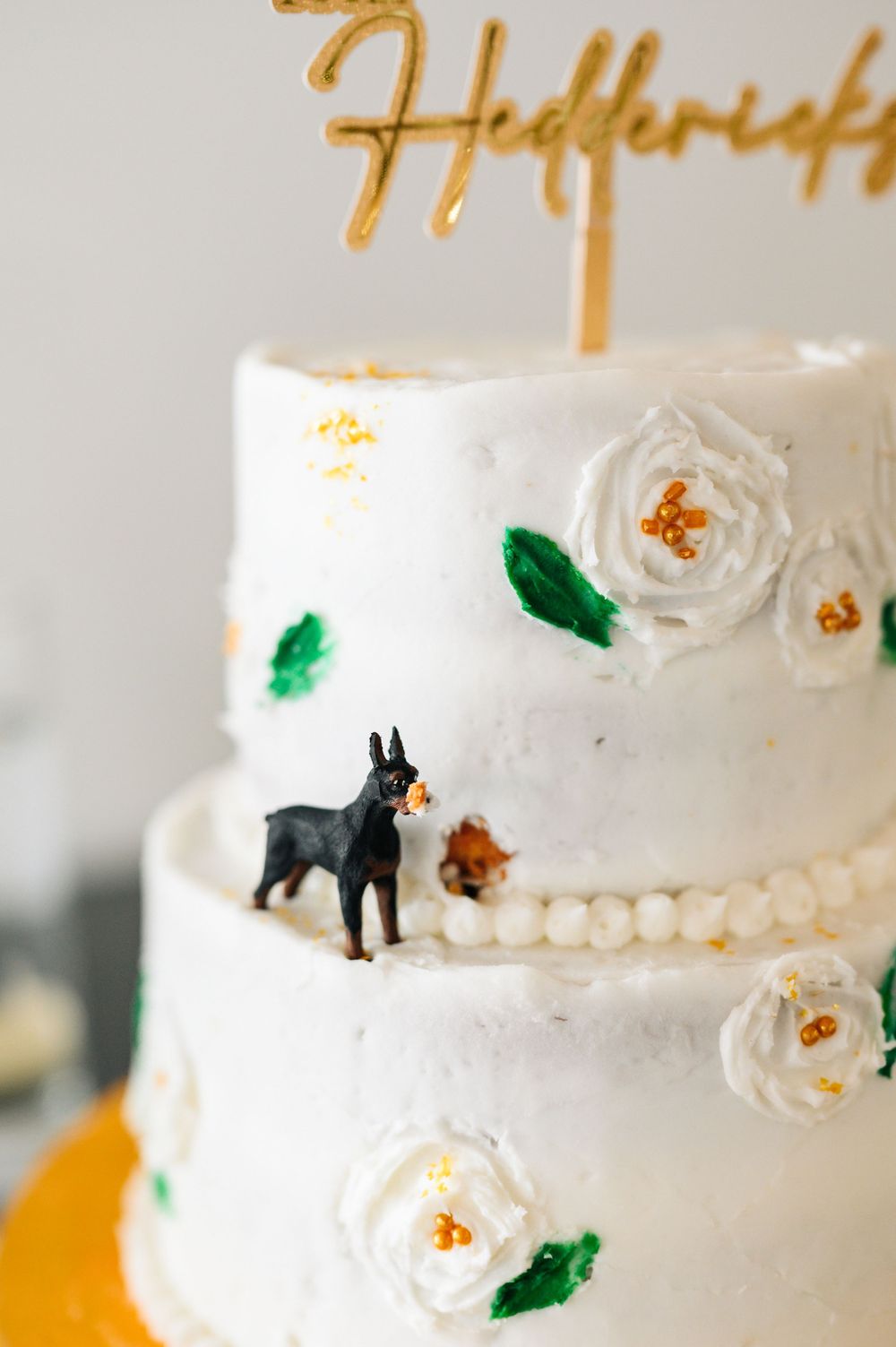 White wedding cake decorated with sugar flowers, gold accents, and a miniature black dog figurine.